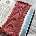 A detailed close-up of a pink knitted bookmark with elegant cable patterns and a scalloped edge. The bookmark is placed on an open book, showcasing its intricate design against the printed text.