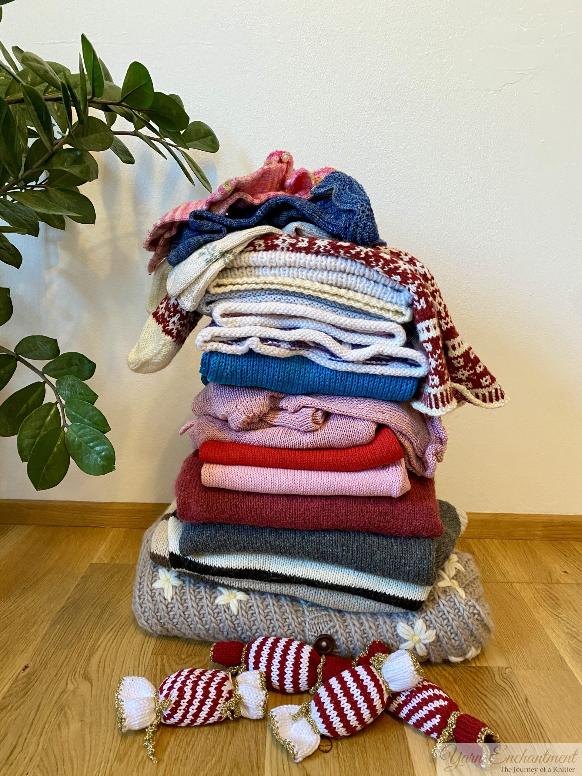 A tall stack of handmade knitted items on a wooden floor, including sweaters, hats, and mittens in red, blue, pink, grey, and white, with a green plant beside them.