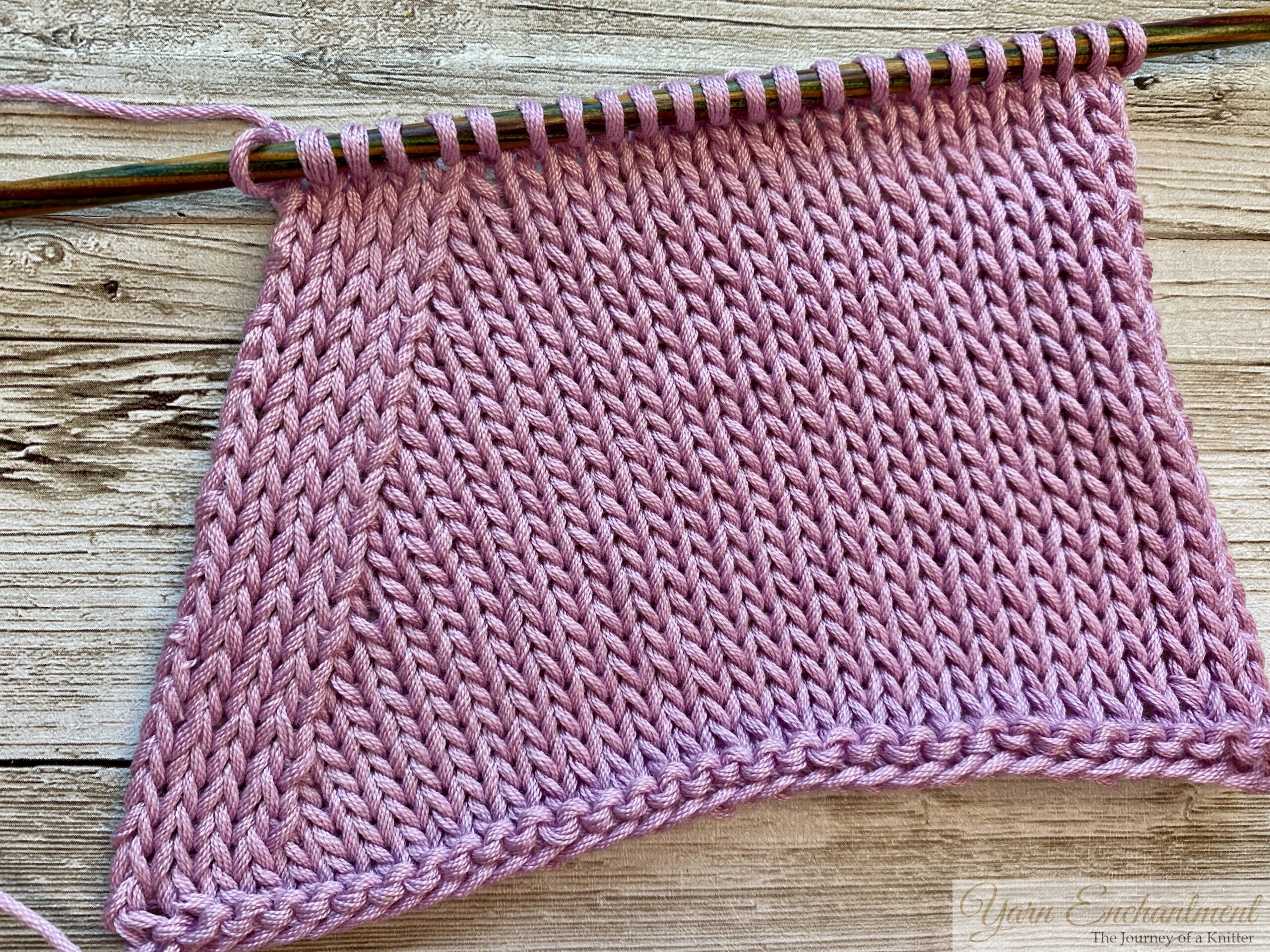 A close-up of a knitted swatch in light pink yarn, showcasing a smooth stockinette stitch pattern with visible shaping created by 'purl 2 together' decreases. The knitting is on wooden needles with a colorful gradient design, placed on a rustic wooden surface.