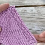 A triangular knitted swatch in light pink yarn, featuring the wrong side with shaping achieved through "purl 2 together" decreases. The piece is on wooden knitting needles with a colorful gradient design, laid on a rustic wooden surface.