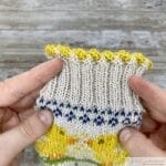 Close-up of hands holding a knitted piece with a completed Latvian Twist edge in yellow yarn. The twist creates a decorative, braided effect at the top of the ribbed section of the knitting.