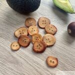 A collection of finished avocado pit buttons displayed on a wooden surface, with a whole avocado, a halved avocado, and an avocado pit in the background.