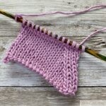 A pink stockinette stitch swatch on colorful wooden knitting needles, resting on a wooden surface.