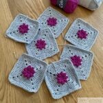 A group of seven crocheted granny squares with pink flowers in the center and light gray borders, neatly arranged on a wooden surface. Two skeins of yarn, one pink and one gray, are placed in the background.