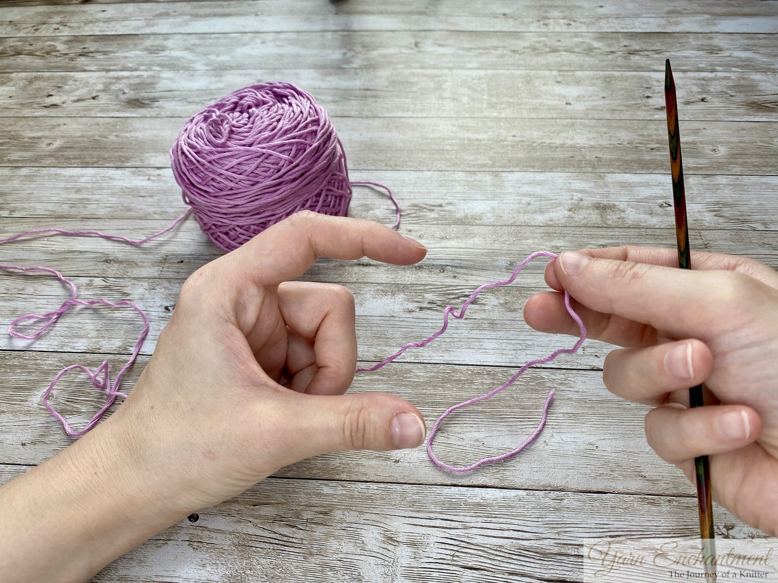 Close-up of hands demonstrating the long tail cast-on technique in knitting. The left hand forms a slingshot shape with the yarn looped around the thumb and index finger, while the right hand holds a knitting needle positioned under the yarn loop on the thumb. A ball of light purple yarn is visible in the background on a wooden surface.
