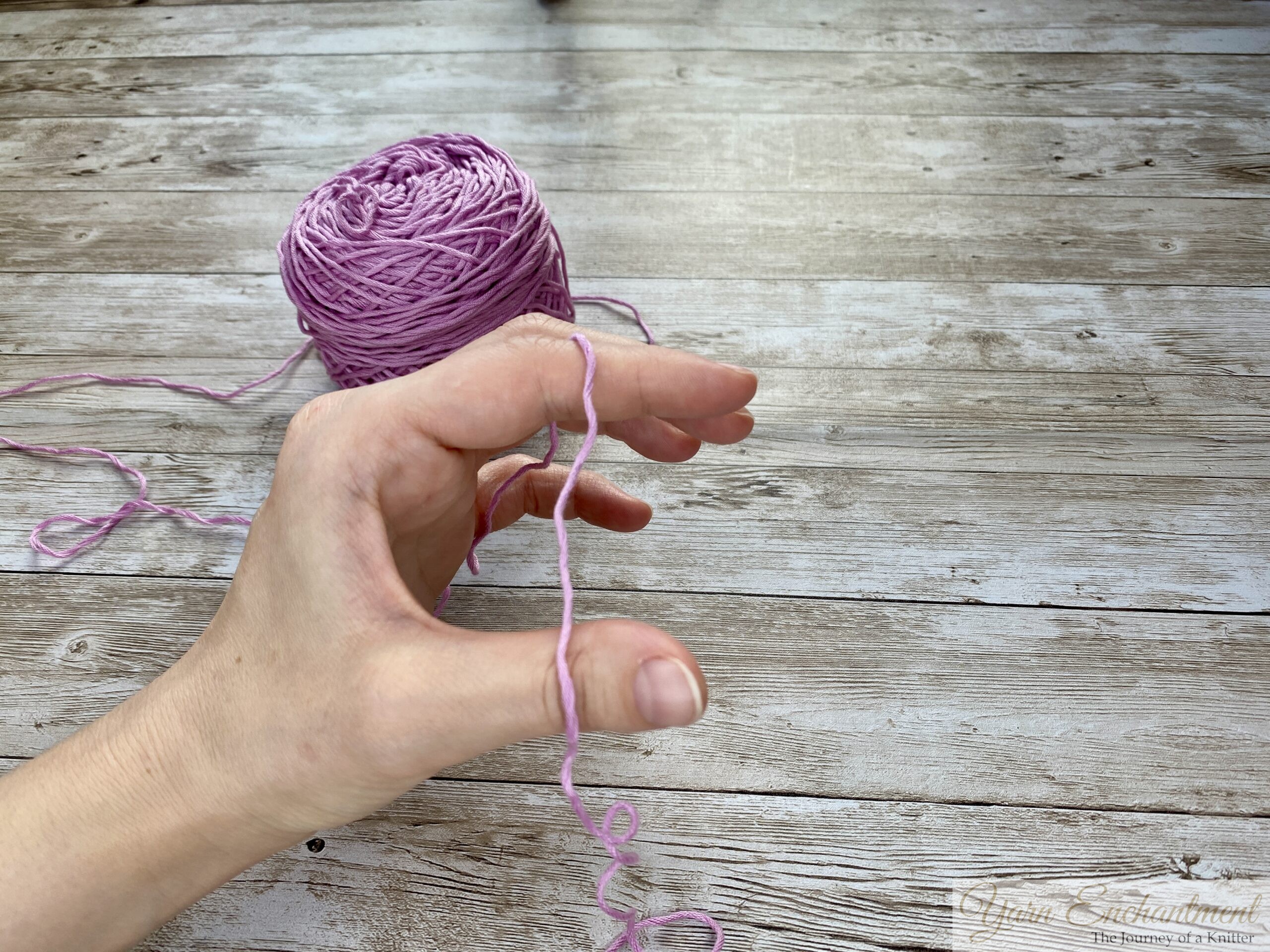 Close-up of hands demonstrating the long tail cast-on technique in knitting. The left hand forms a slingshot shape with the yarn looped around the thumb and index finger, while the right hand holds a knitting needle positioned under the yarn loop on the thumb. A ball of light purple yarn is visible in the background on a wooden surface.