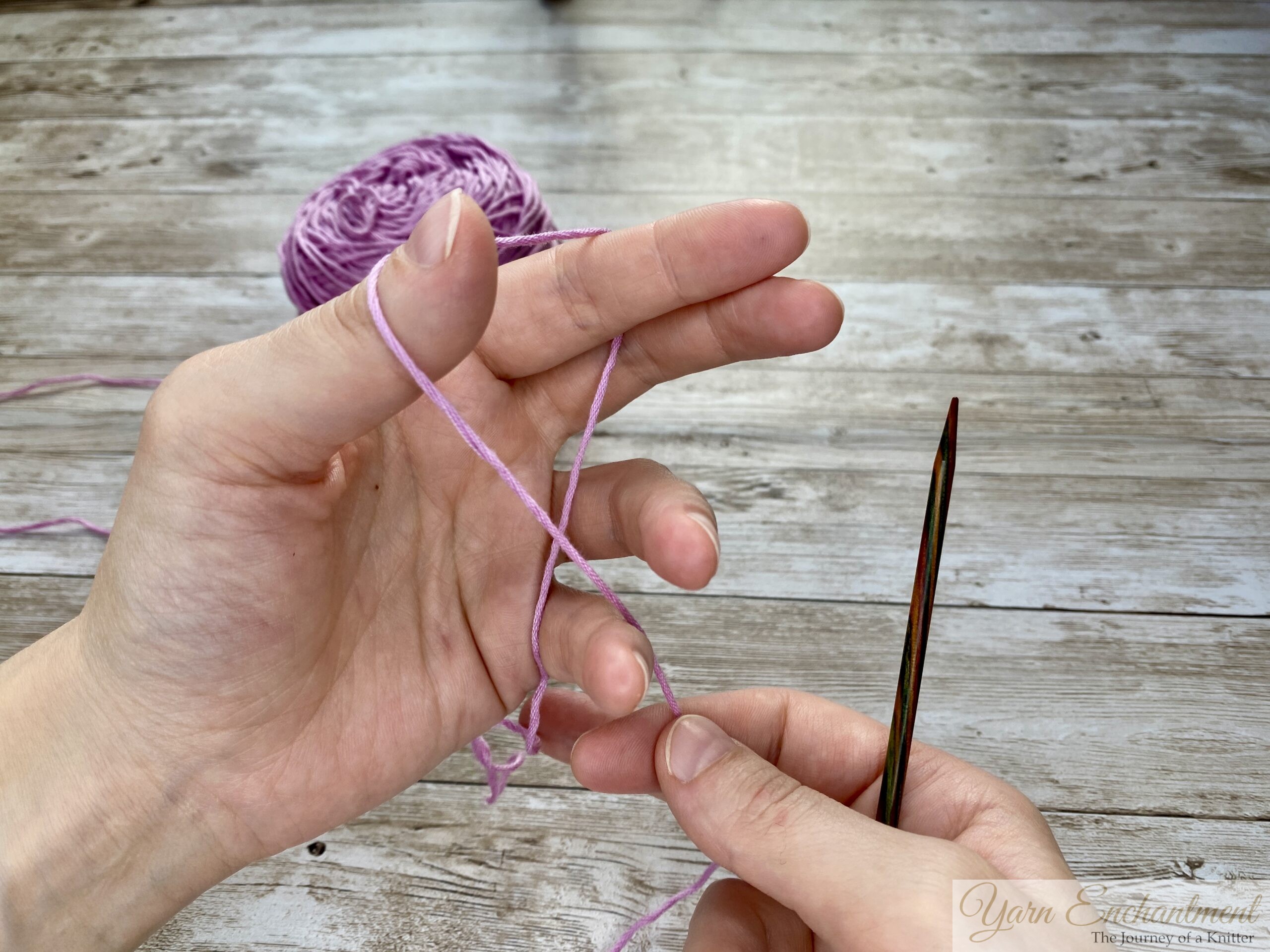 Close-up of hands demonstrating the long tail cast-on technique in knitting. The left hand forms a slingshot shape with the yarn looped around the thumb and index finger, while the right hand holds a knitting needle positioned under the yarn loop on the thumb. A ball of light purple yarn is visible in the background on a wooden surface.