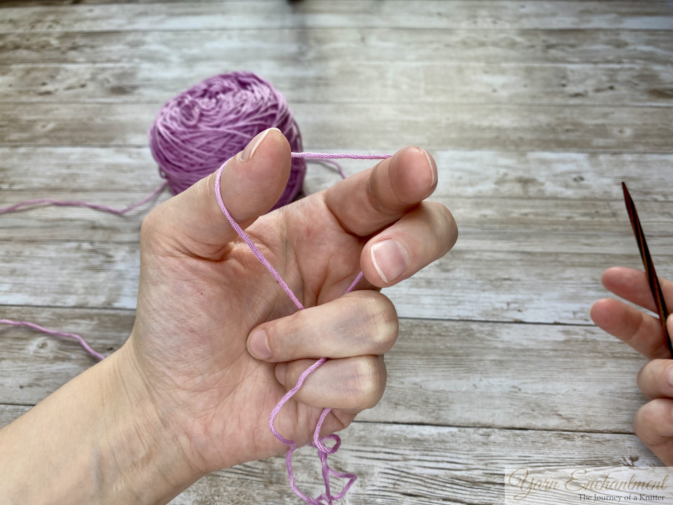Close-up of hands demonstrating the long tail cast-on technique in knitting. The left hand forms a slingshot shape with the yarn looped around the thumb and index finger, while the right hand holds a knitting needle positioned under the yarn loop on the thumb. A ball of light purple yarn is visible in the background on a wooden surface.