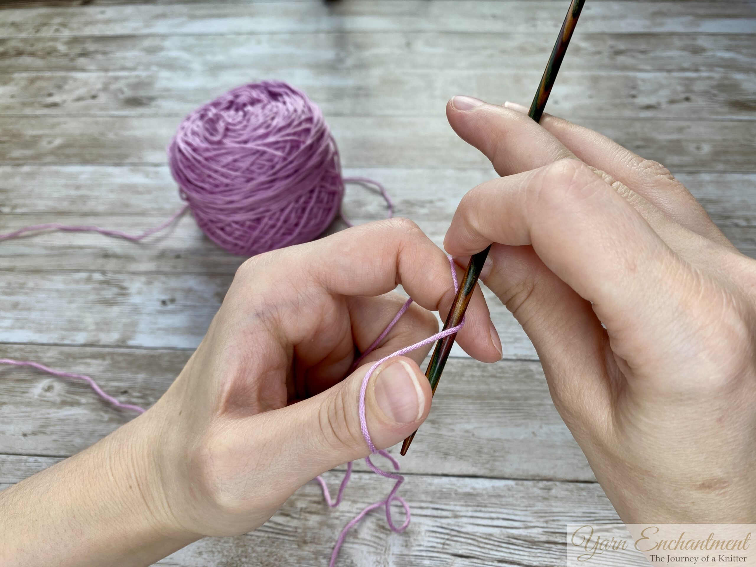 Close-up of hands demonstrating the long tail cast-on technique in knitting. The left hand forms a slingshot shape with the yarn looped around the thumb and index finger, while the right hand holds a knitting needle positioned under the yarn loop on the thumb. A ball of light purple yarn is visible in the background on a wooden surface.