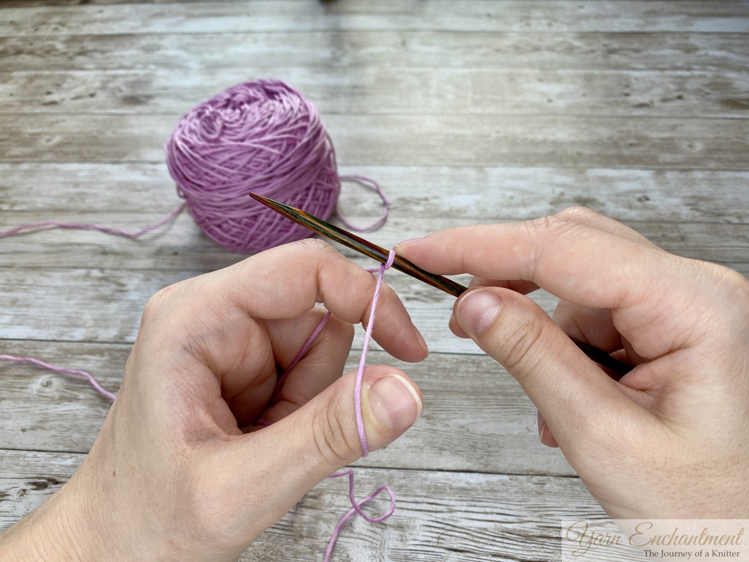 Close-up of hands demonstrating the long tail cast-on technique in knitting. The left hand forms a slingshot shape with the yarn looped around the thumb and index finger, while the right hand holds a knitting needle positioned under the yarn loop on the thumb. A ball of light purple yarn is visible in the background on a wooden surface.