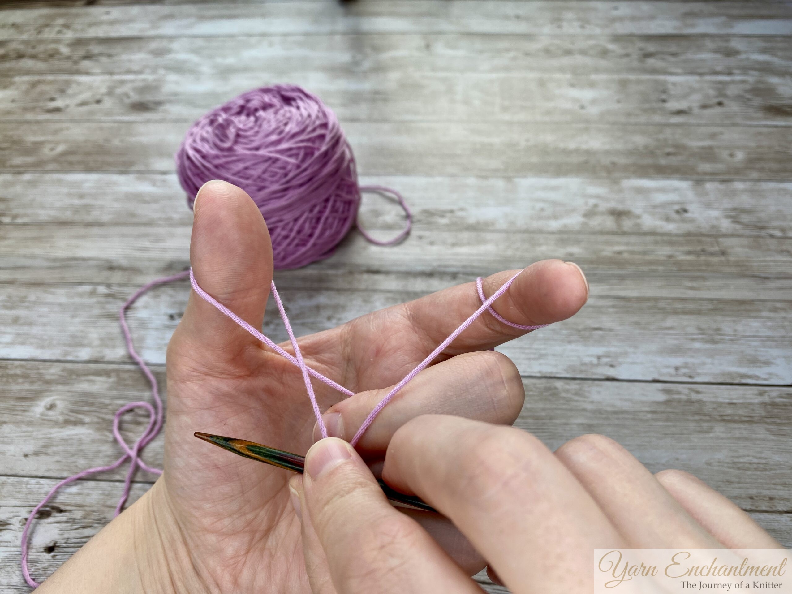Close-up of hands demonstrating the long tail cast-on technique in knitting. The left hand forms a slingshot shape with the yarn looped around the thumb and index finger, while the right hand holds a knitting needle positioned under the yarn loop on the thumb. A ball of light purple yarn is visible in the background on a wooden surface.