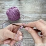 Close-up of hands demonstrating the long tail cast-on technique in knitting. The left hand forms a slingshot shape with the yarn looped around the thumb and index finger, while the right hand holds a knitting needle positioned under the yarn loop on the thumb. A ball of light purple yarn is visible in the background on a wooden surface.