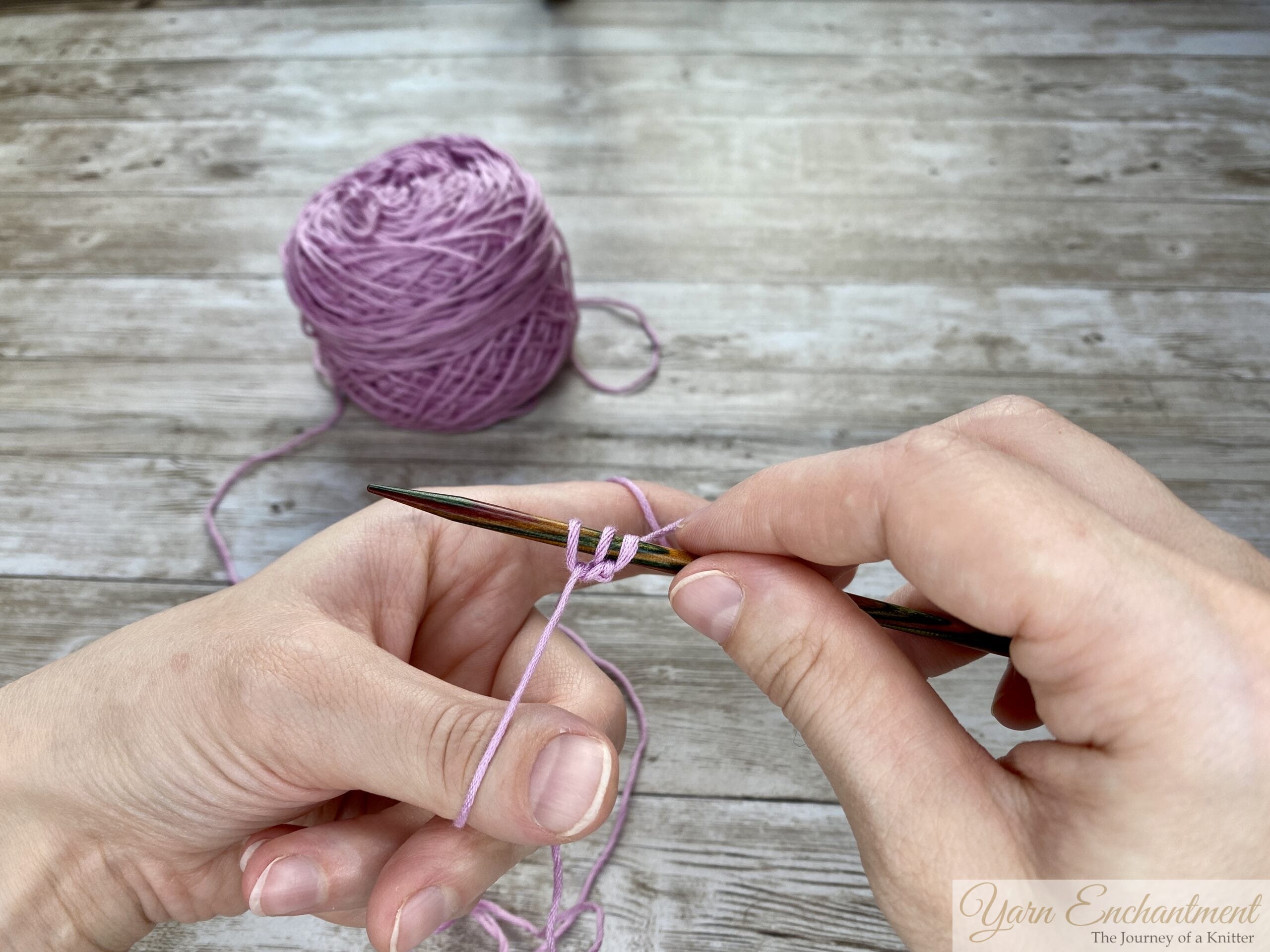 Close-up of hands demonstrating the long tail cast-on technique in knitting, with a pink yarn.