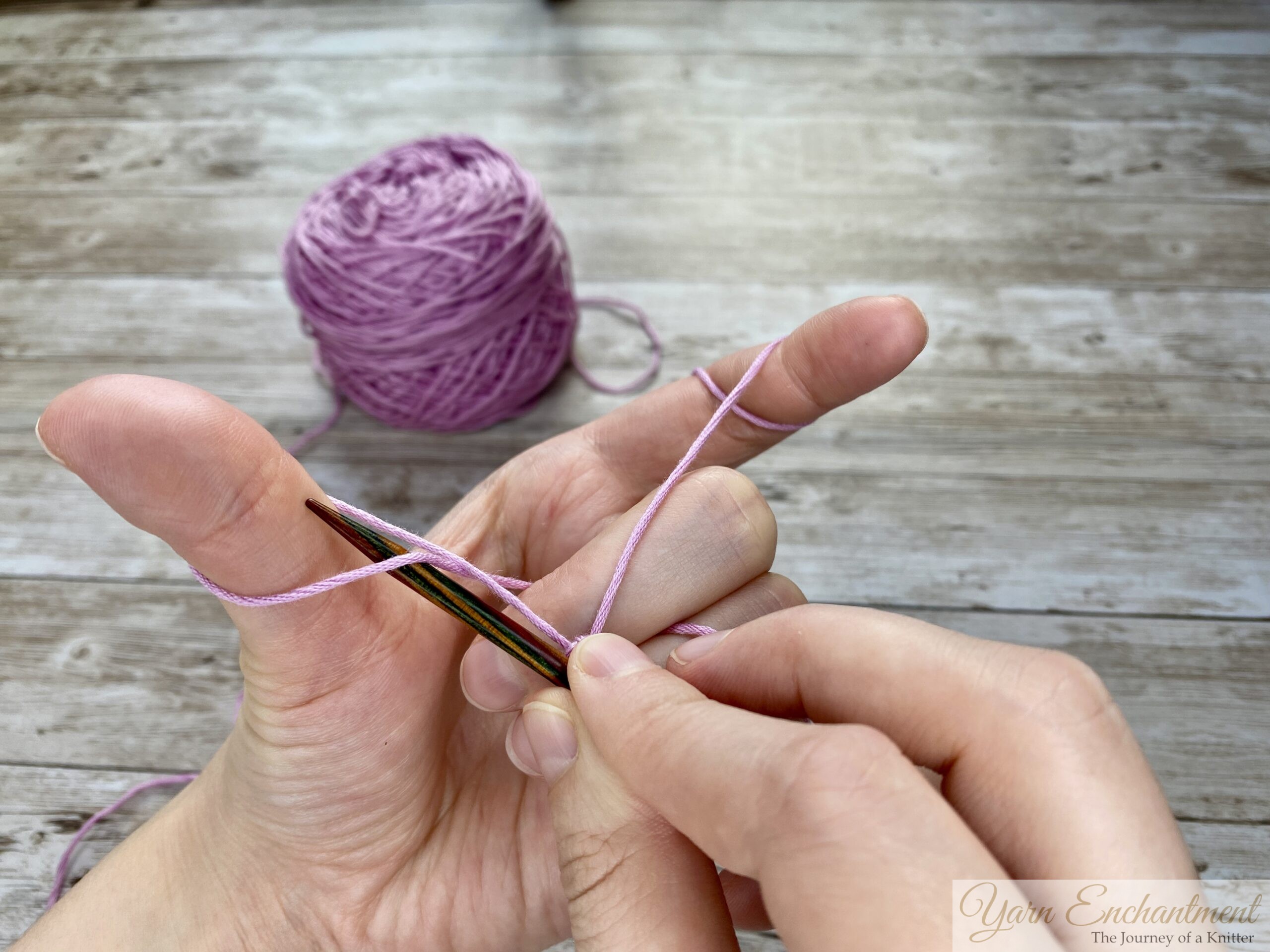 Close-up of hands demonstrating the long tail cast-on technique in knitting. The left hand forms a slingshot shape with the yarn looped around the thumb and index finger, while the right hand holds a knitting needle positioned under the yarn loop on the thumb. A ball of light purple yarn is visible in the background on a wooden surface.