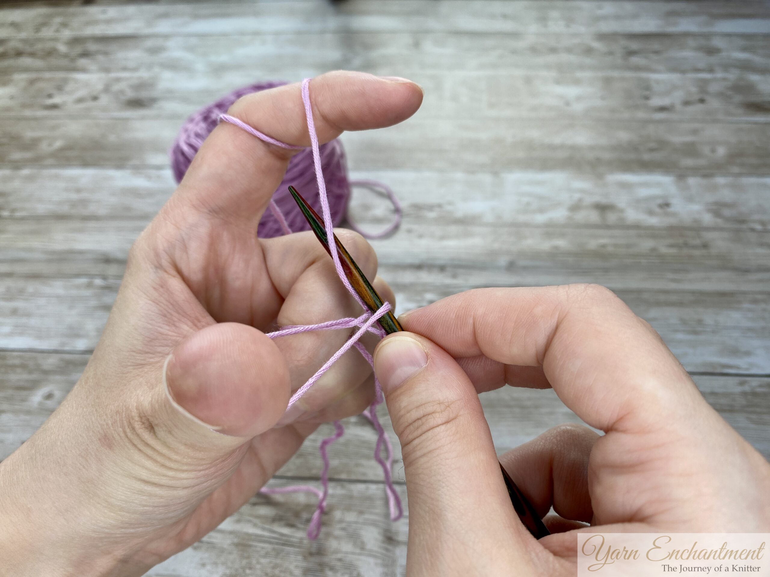 Close-up of hands demonstrating the long tail cast-on technique in knitting. The left hand forms a slingshot shape with the yarn looped around the thumb and index finger, while the right hand holds a knitting needle positioned under the yarn loop on the thumb. A ball of light purple yarn is visible in the background on a wooden surface