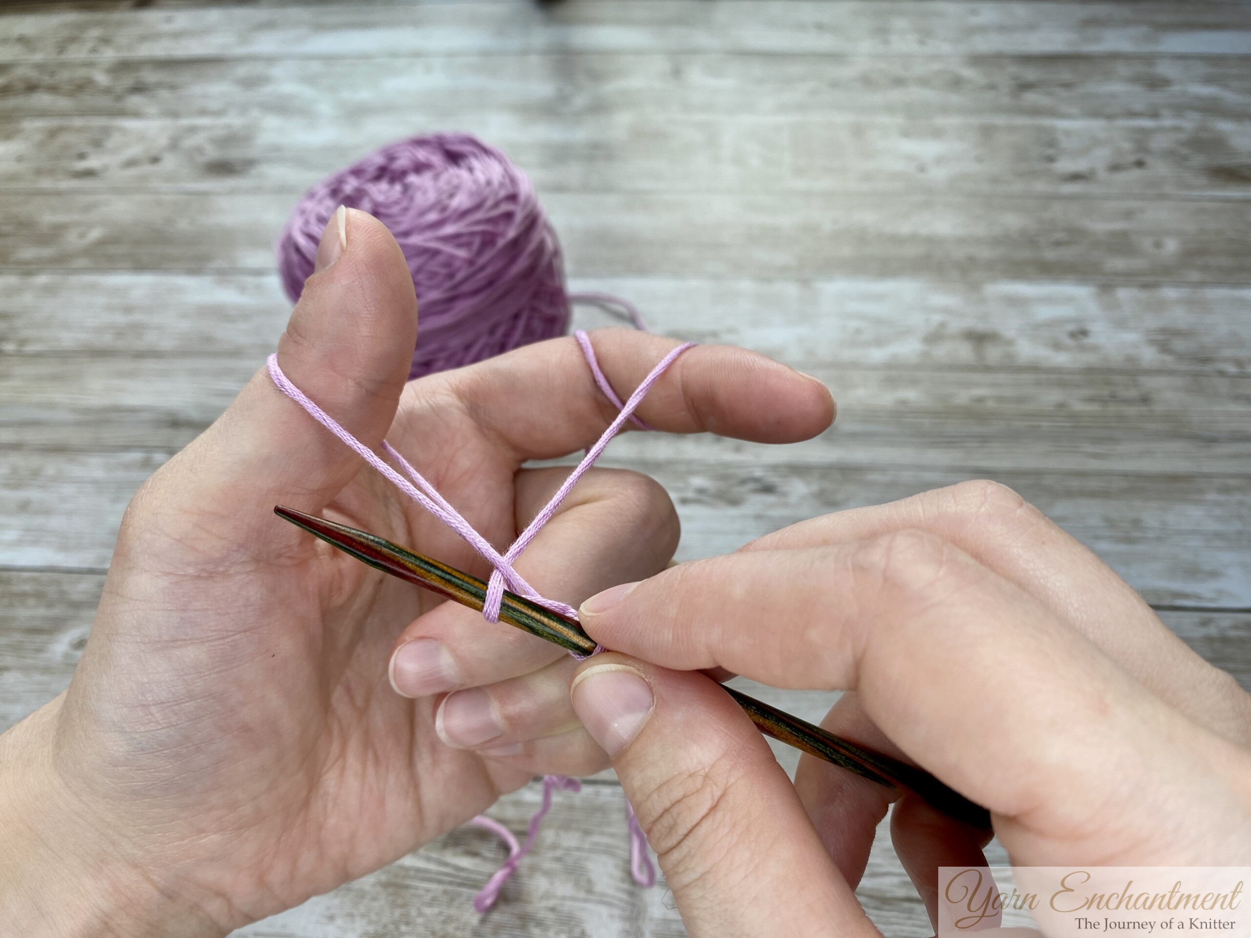 Close-up of hands demonstrating the long tail cast-on technique in knitting. The left hand forms a slingshot shape with the yarn looped around the thumb and index finger, while the right hand holds a knitting needle positioned under the yarn loop on the thumb. A ball of light purple yarn is visible in the background on a wooden surface