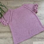Full view of the completed light pink knitted shirt with short ruffled sleeves. The shirt is displayed flat on a wooden background, and both the decorative raglan details and the ruffled sleeve edges are visible.