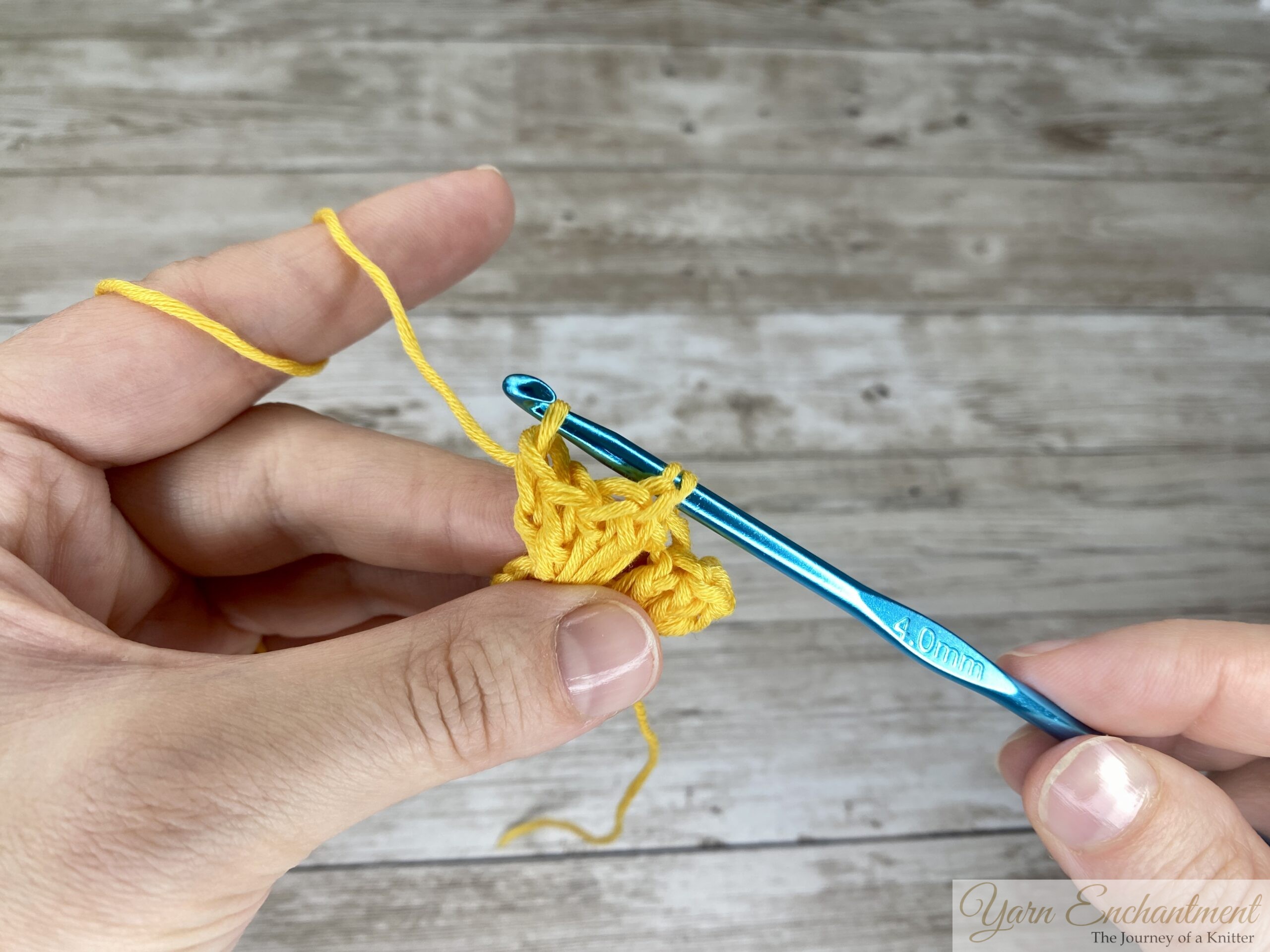 Close-up of hands crocheting with bright yellow yarn, using a 4.0 mm hook to pull a loop through stitches. The left hand holds the growing crochet piece while guiding tension with the index finger.