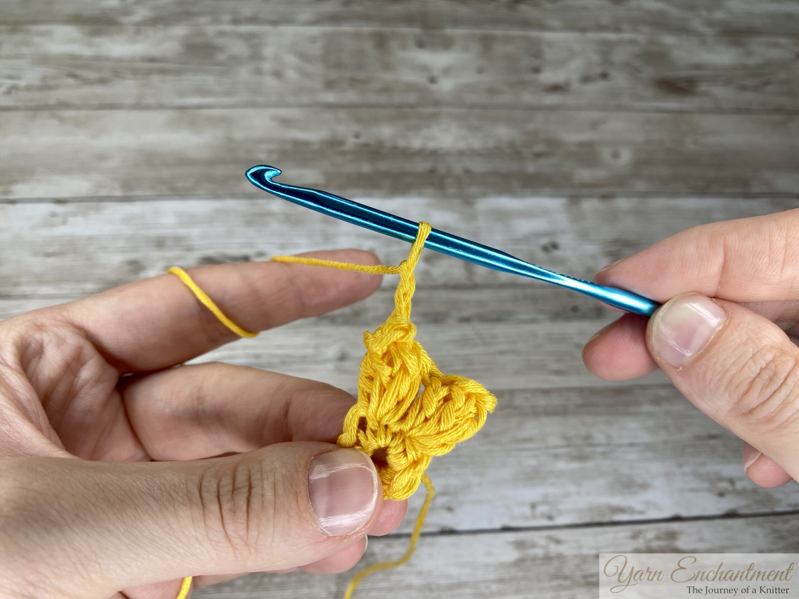 Close-up of crochet hook completing two chain stitches with yellow yarn, creating space between 3D granny square clusters.