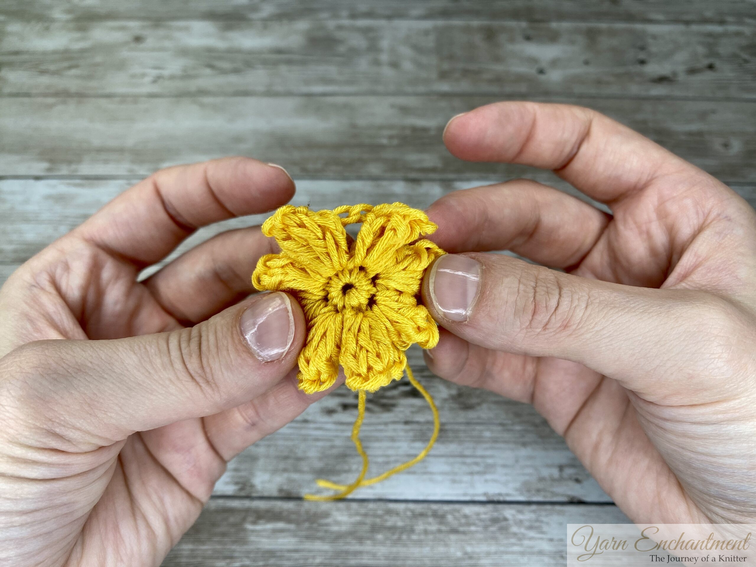 A person holding a vibrant yellow crochet flower between their hands, resting on a light wooden surface