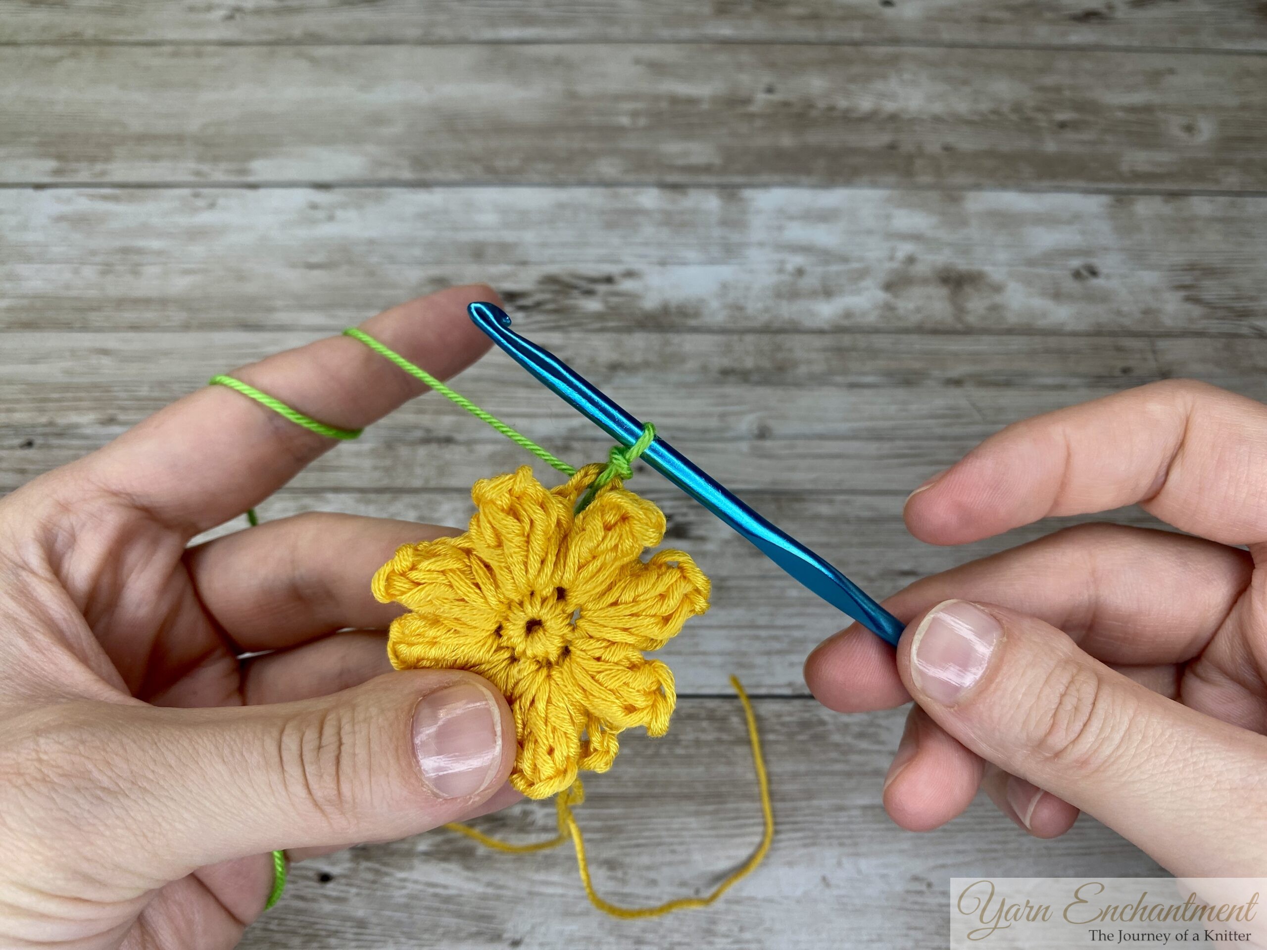 A person crocheting a yellow flower with a 3D design, using a blue crochet hook and green yarn to begin the next round. One hand holds the flower, while the other guides the hook, with a wooden background underneath.