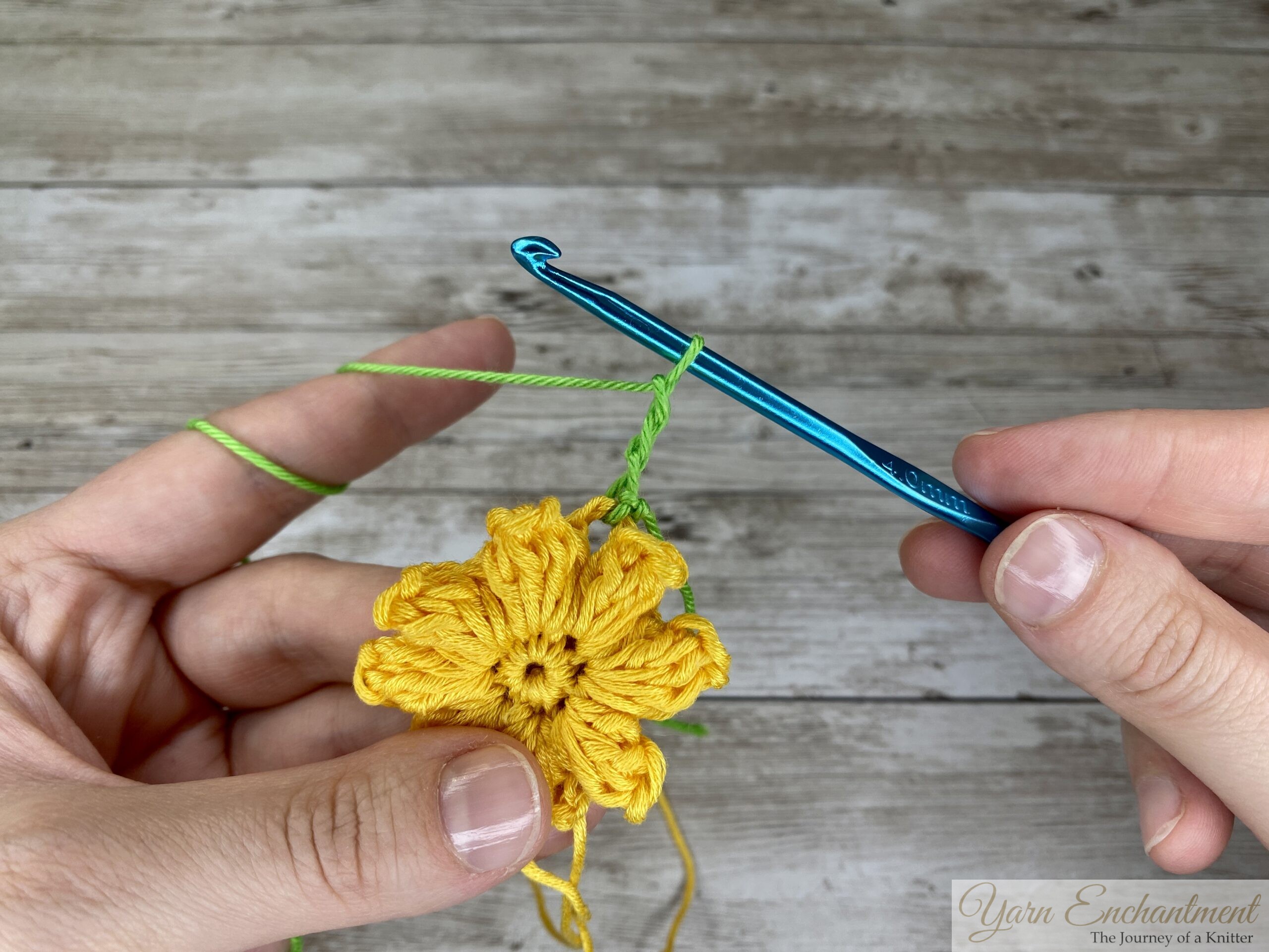 A close-up of a crochet project showing a yellow 3D flower being worked on, with a green yarn loop on a blue crochet hook. The hands are in the process of creating chain stitches.