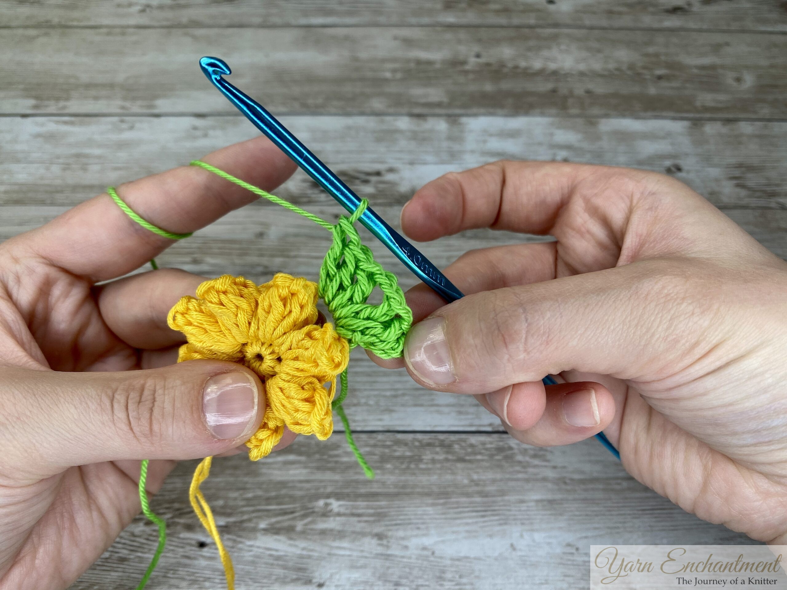 A crochet project showing progress of working the first corner from a granny square border with green yarn. The corner is worked into the chain spaces from the yellow 3D flower. A blue crochet hook is held in one hand, while the other holds the flower.