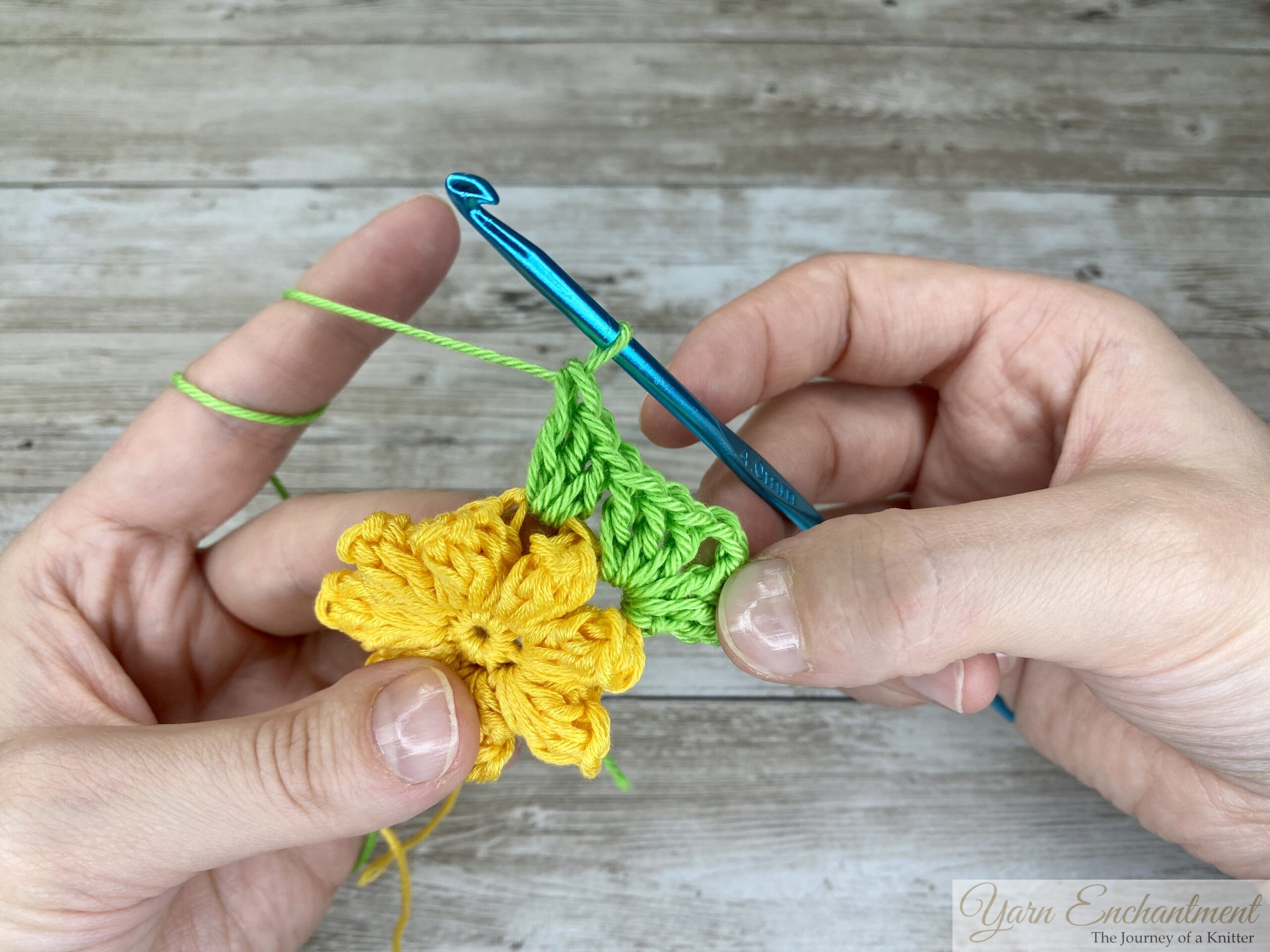 A detailed view of a yellow crochet flower with green stitches forming a granny square base. A blue crochet hook is positioned in the green yarn, with both hands guiding the project.