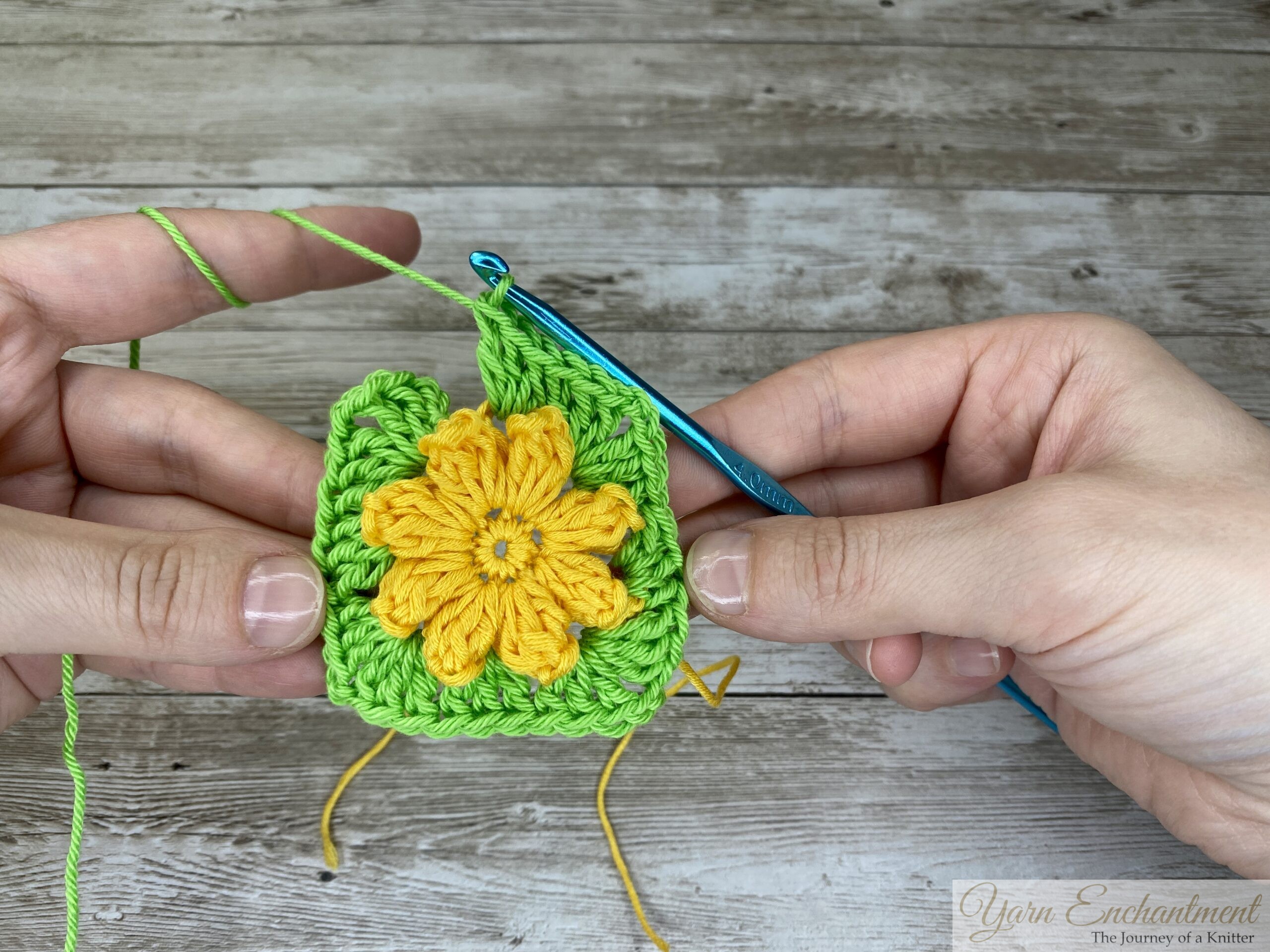A crochet project showing a completed yellow 3D flower at the center of a bright green granny square in progress. A blue crochet hook is inserted into the green yarn, with both hands holding the project on a wooden surface.