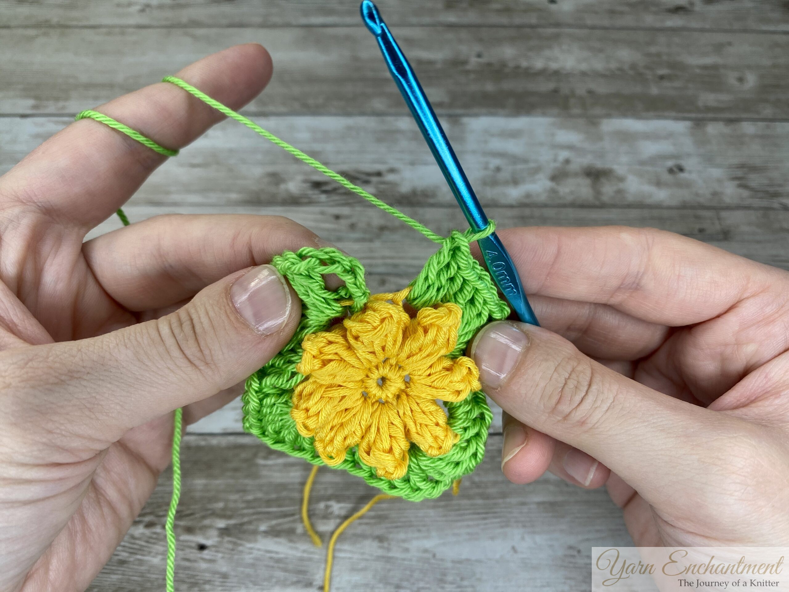 A close-up of a crochet project showing a yellow 3D flower in progress, surrounded by a green granny square base. A blue crochet hook is inserted into the green yarn, with both hands holding the piece.