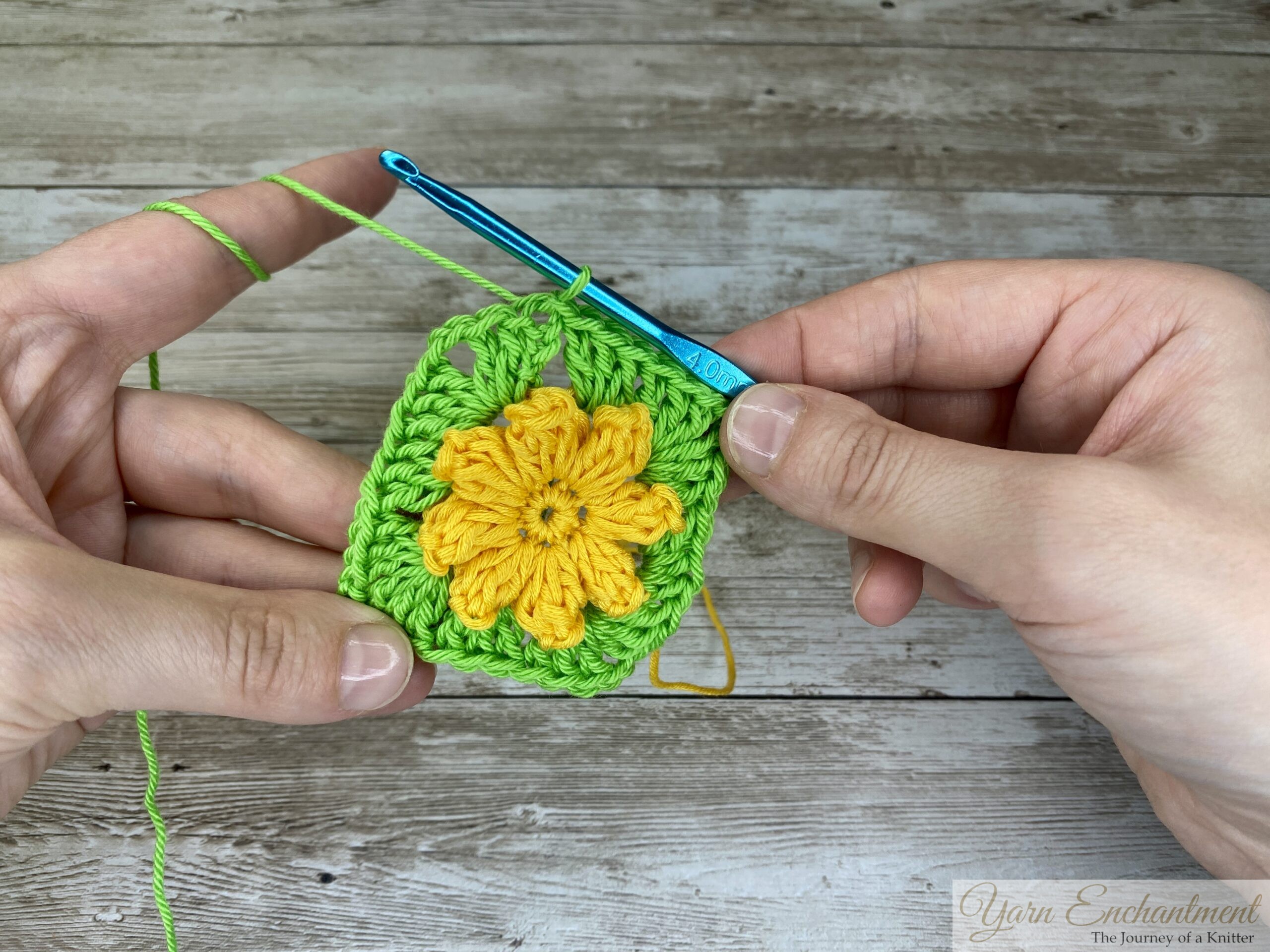 A yellow 3D crochet flower surrounded by a green granny square base, with a blue crochet hook continuing the next round. The project is held by both hands on a wooden surface.