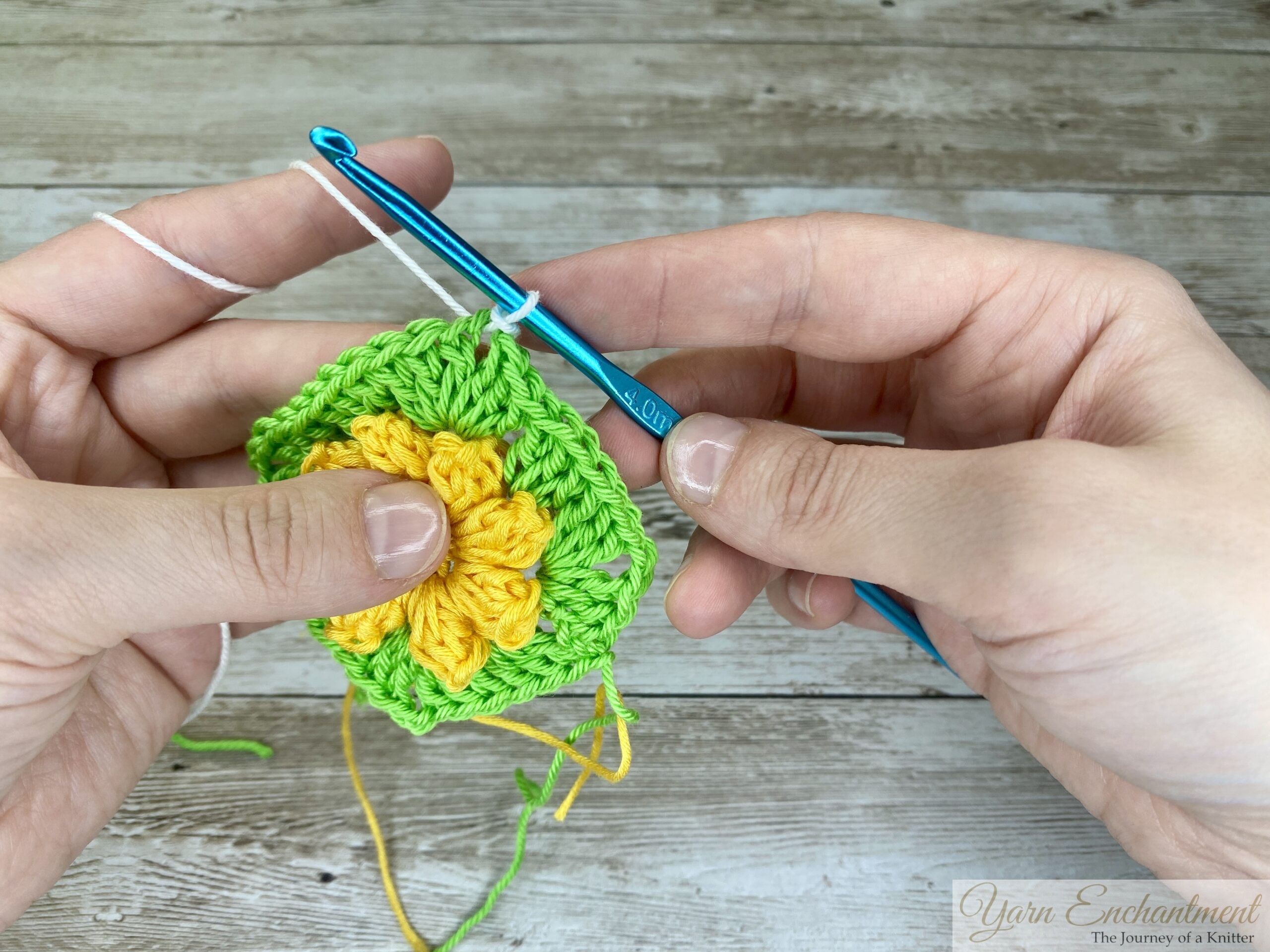 A crochet project showing a yellow 3D flower encased in a green granny square base. A new white yarn is being added with a blue crochet hook, held by both hands.