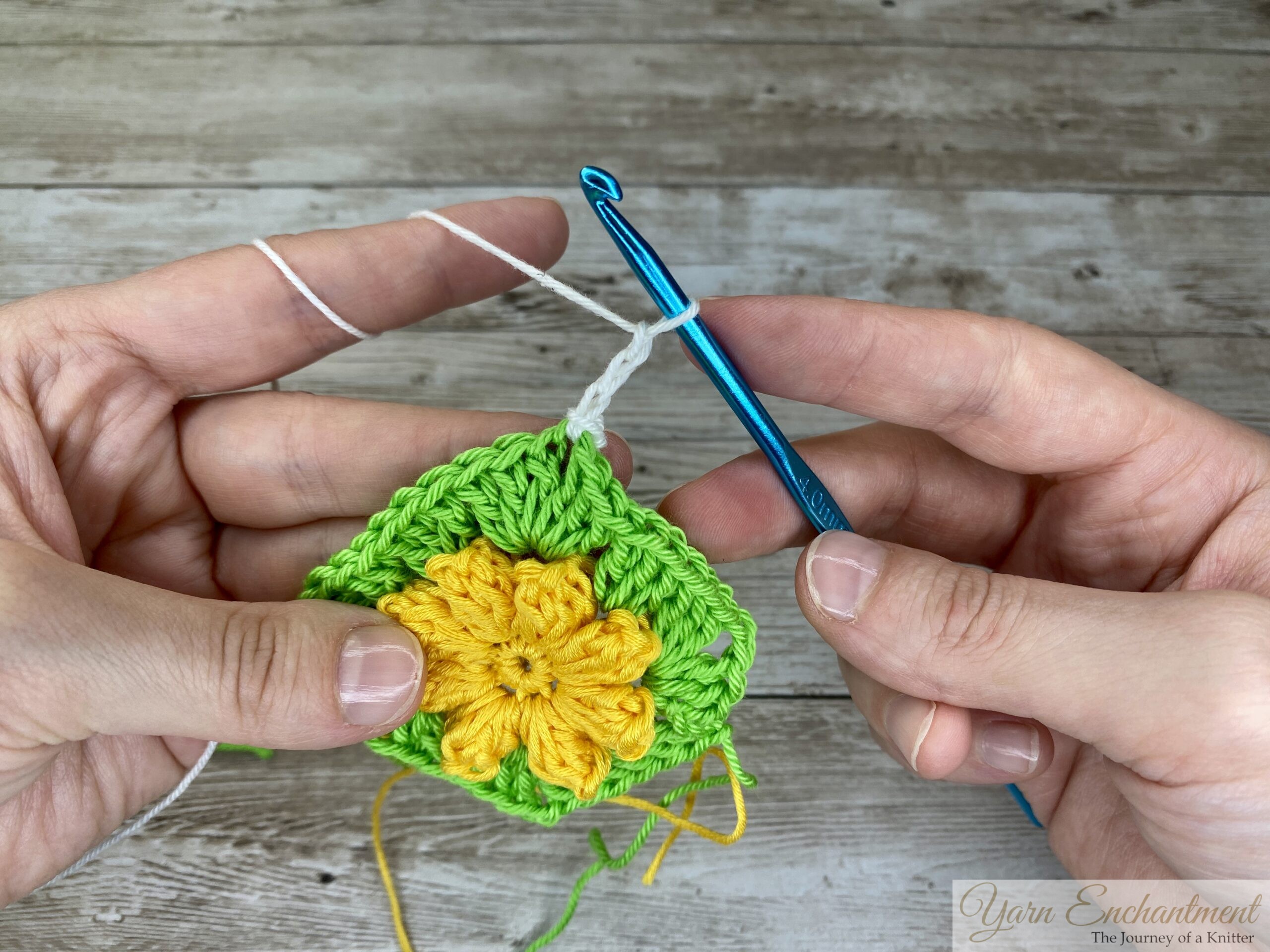 A crochet project in progress, featuring a yellow 3D flower in the center of a green granny square. White yarn has been added, and a blue crochet hook is shown with the first chain stitches.