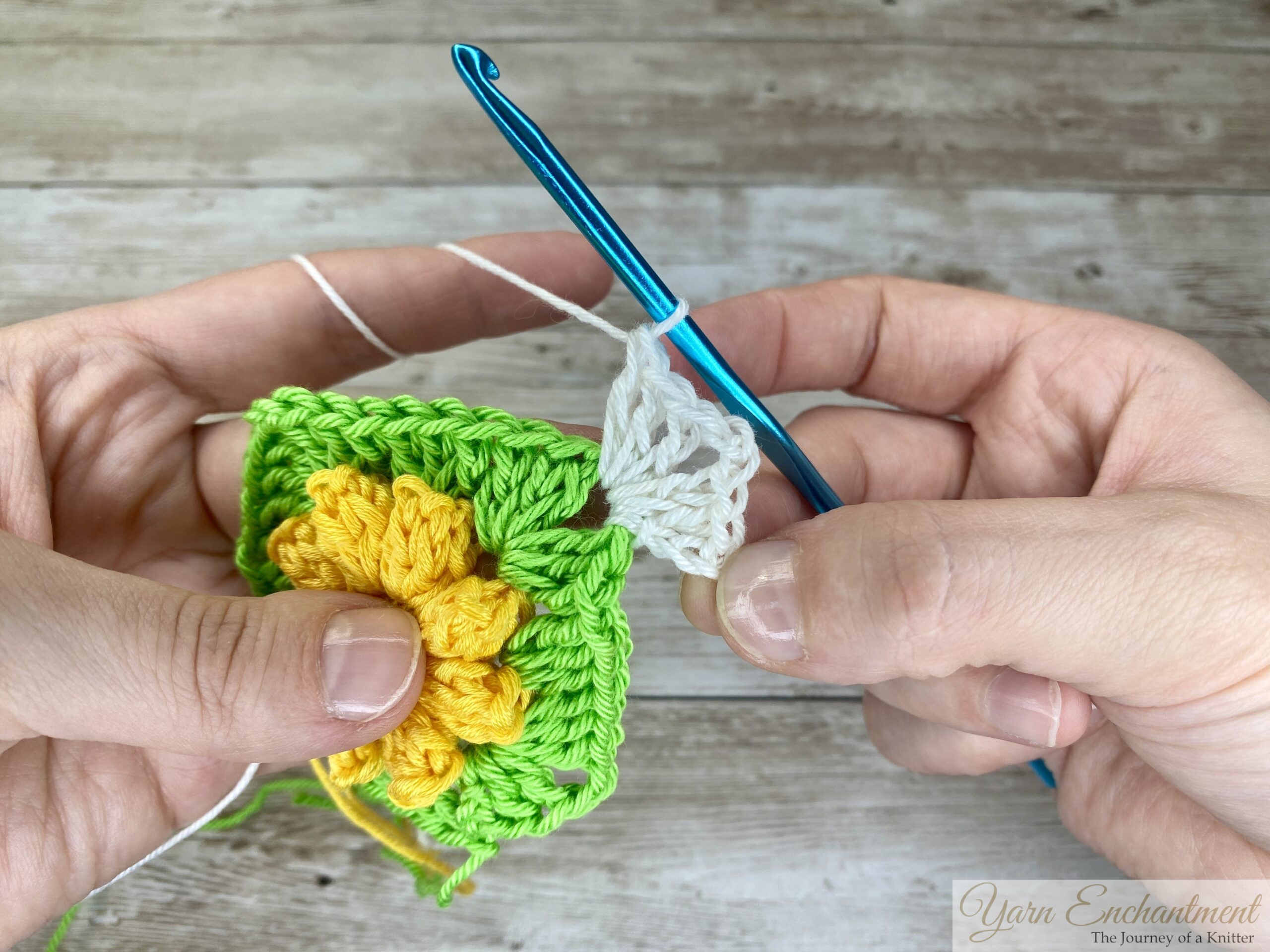 The crochet project showing the yellow flower and green granny square now surrounded by white yarn. A blue crochet hook is working on a double crochet cluster.