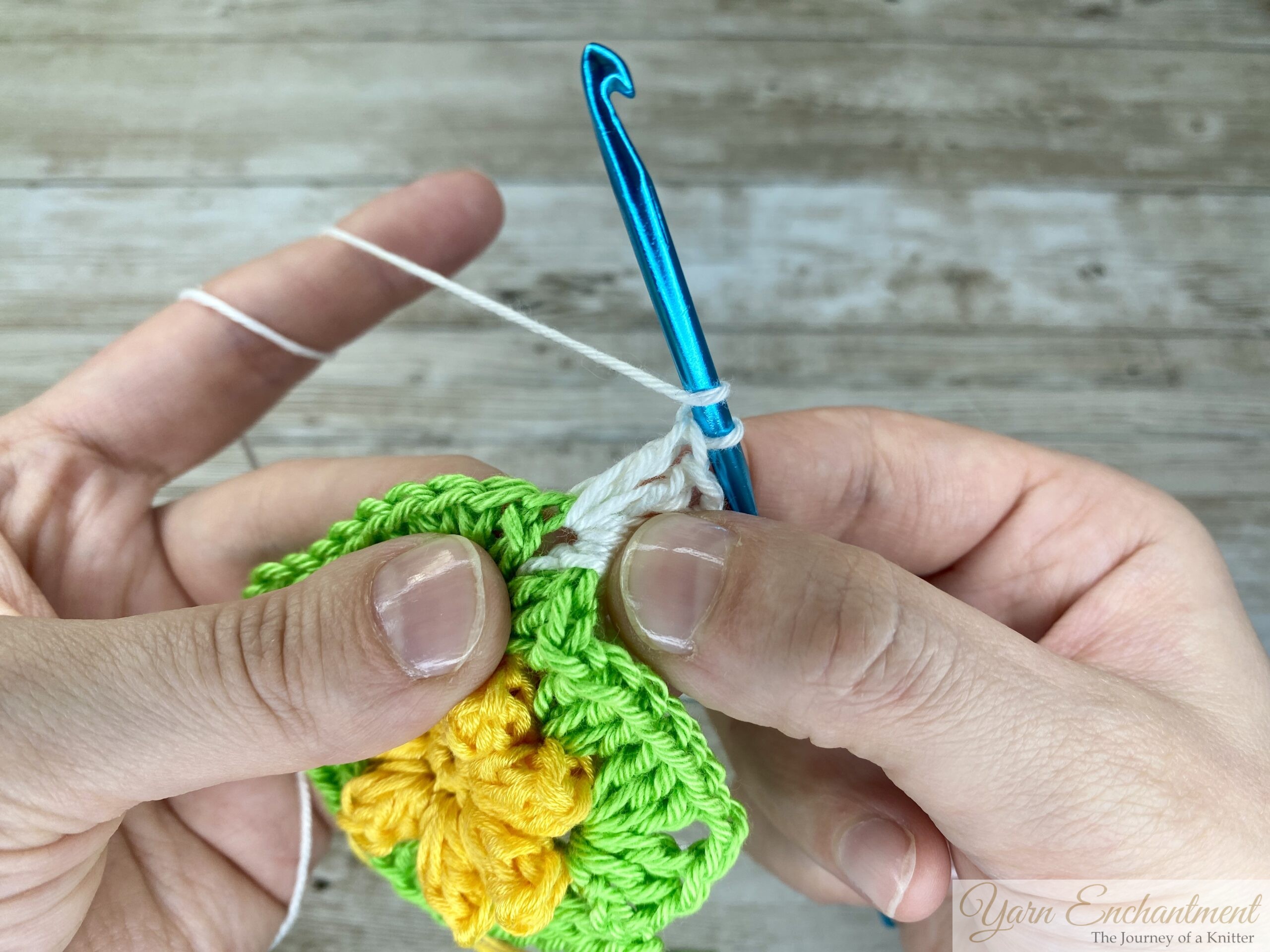 Close-up of a crochet project with a yellow flower and green granny square, as white yarn is being worked into the next stitch with a blue crochet hook.