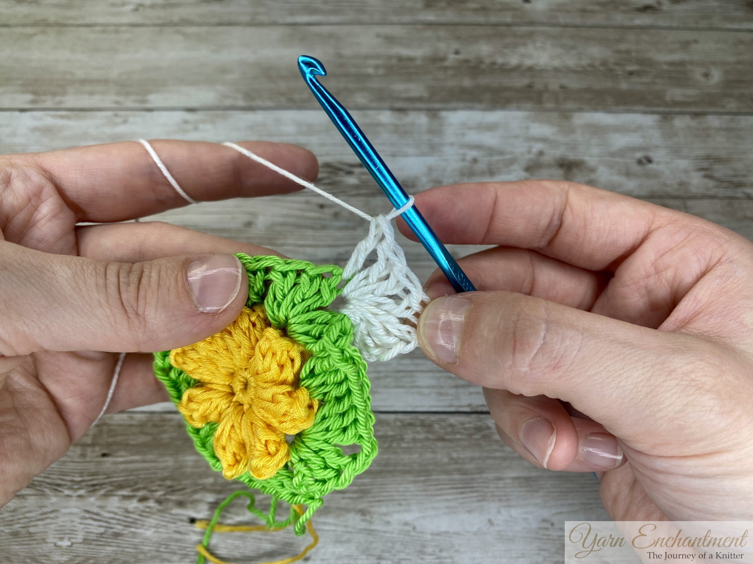 The crochet project progresses with white yarn forming a row of stitches around the green granny square and yellow flower. A blue crochet hook is continuing the stitch.