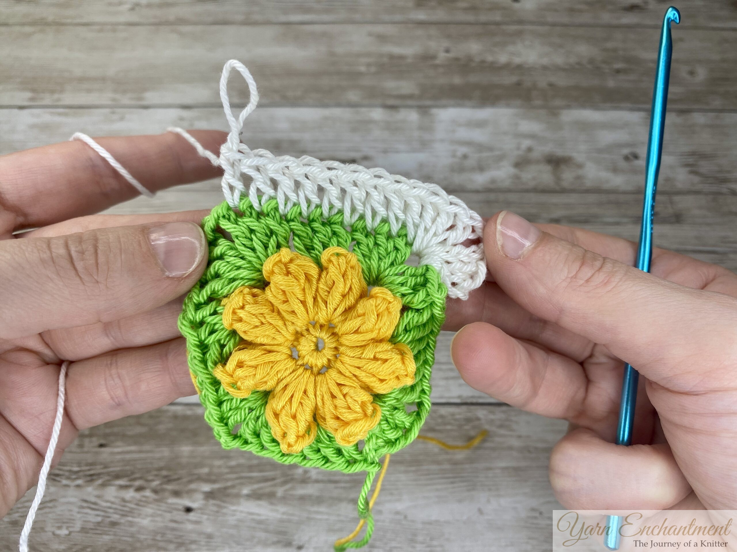 A nearly completed crochet project showing a yellow flower surrounded by green and white yarn. The white border is almost finished, with a blue crochet hook resting on the project.