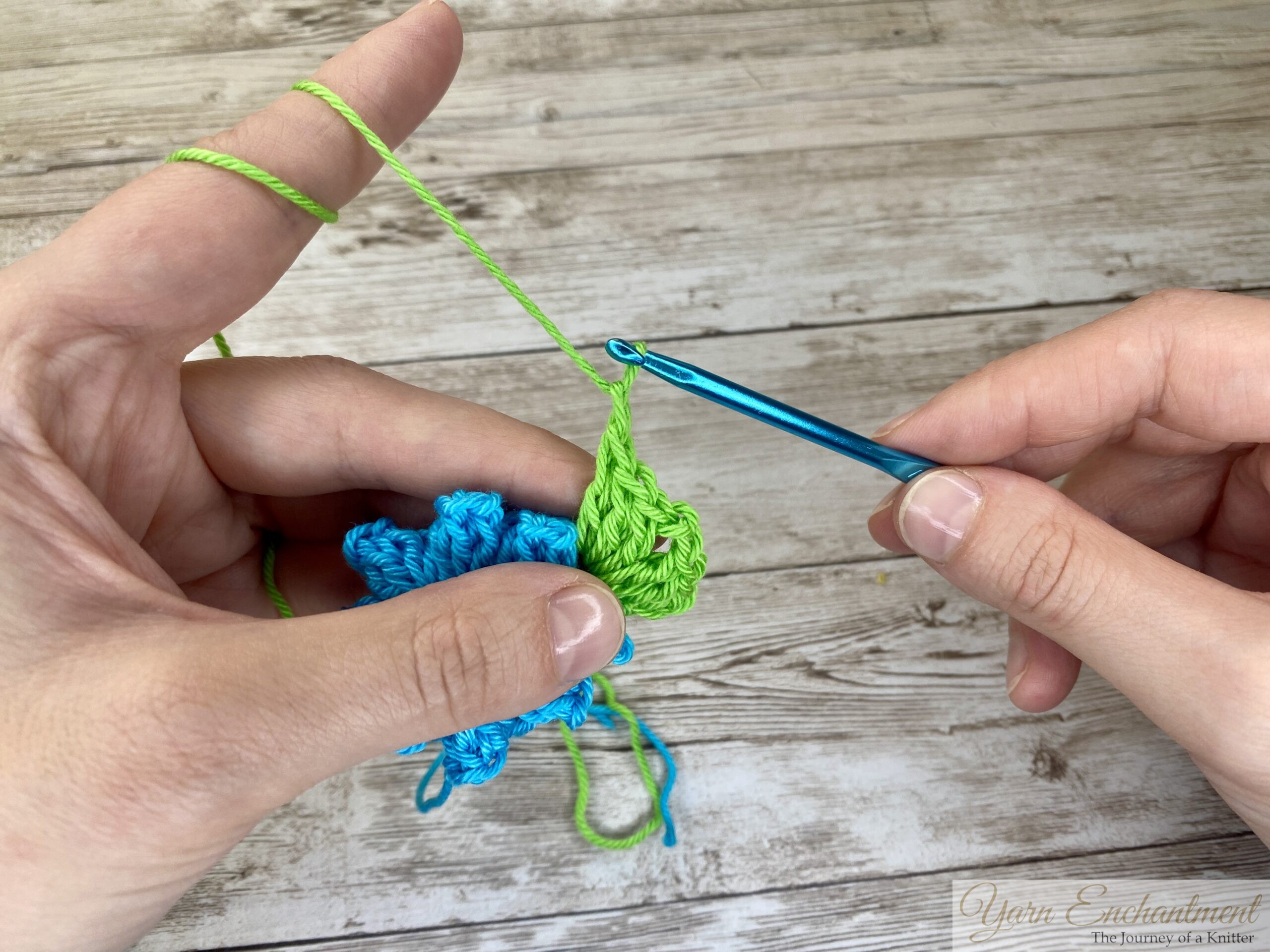 Close-up of a crochet project showing a blue flower centerpiece as green yarn is being looped to form a granny square with a blue crochet hook.