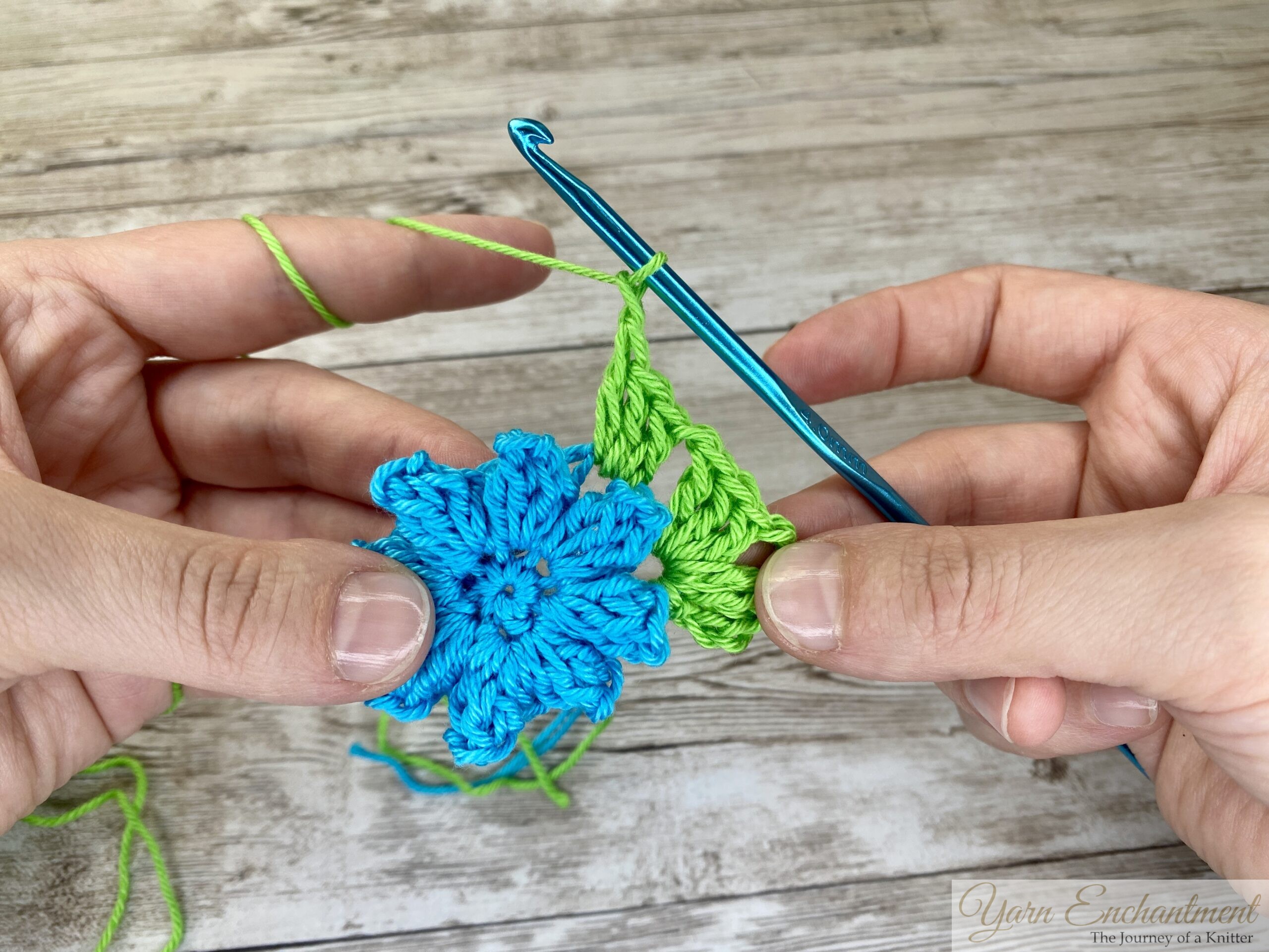 A crochet project showing a blue flower centerpiece with green yarn forming the beginnings of a granny square. A blue crochet hook is in use as the project progresses.