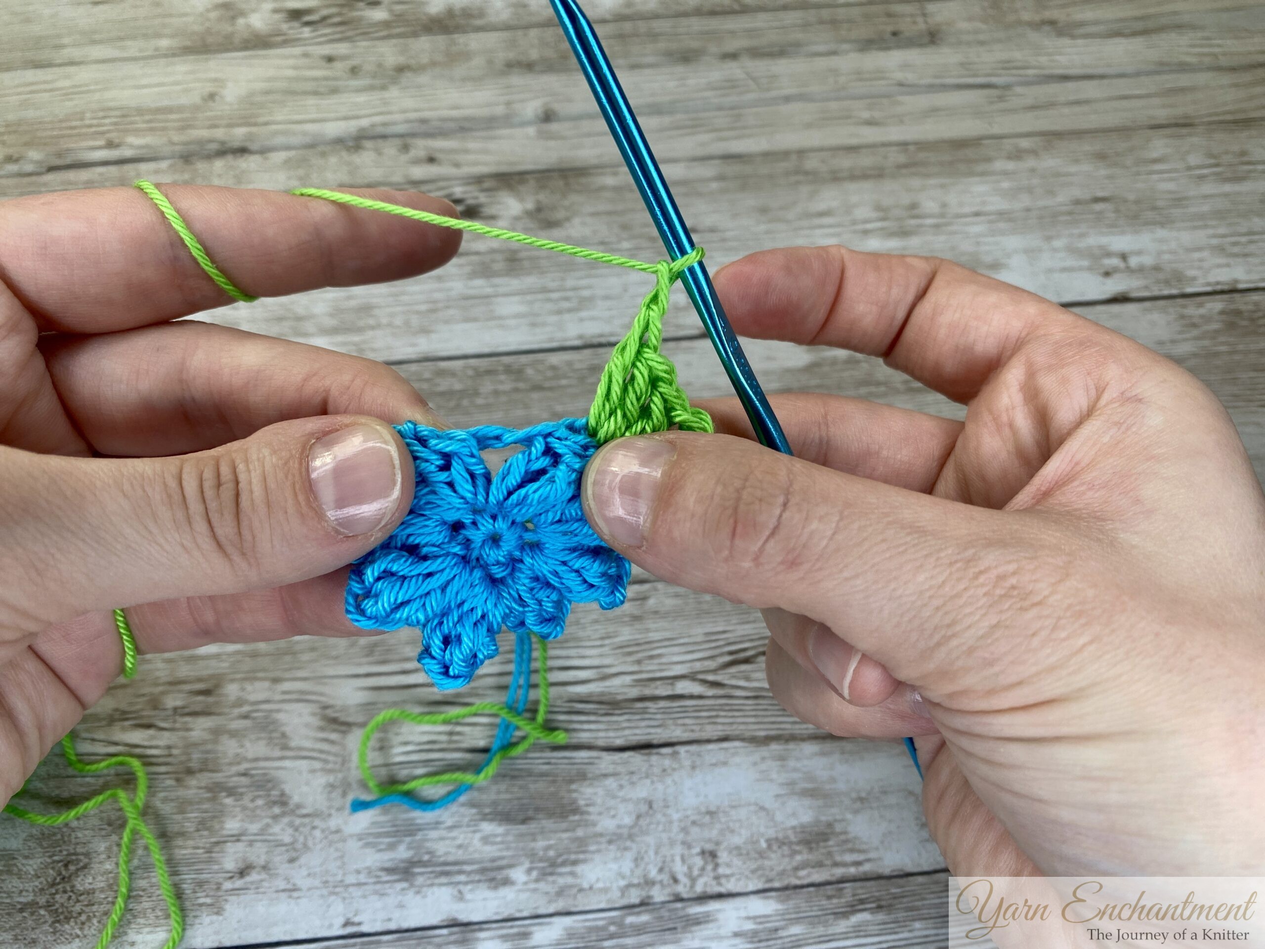 A crochet project in progress showing a bright blue flower centerpiece with green yarn being added in a granny square pattern. A blue crochet hook is actively working on the green stitches.