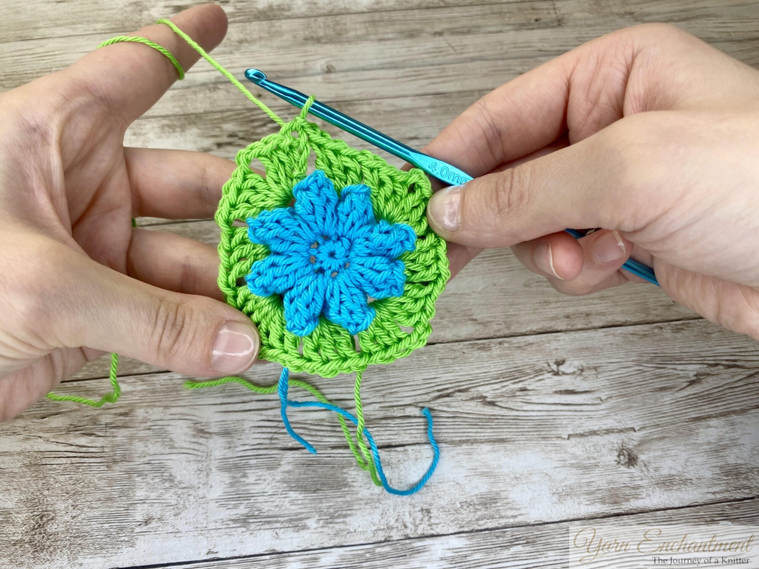 A crochet project showing a blue flower centerpiece surrounded by green yarn forming a granny square. A blue crochet hook is continuing the pattern as both hands hold the project.
