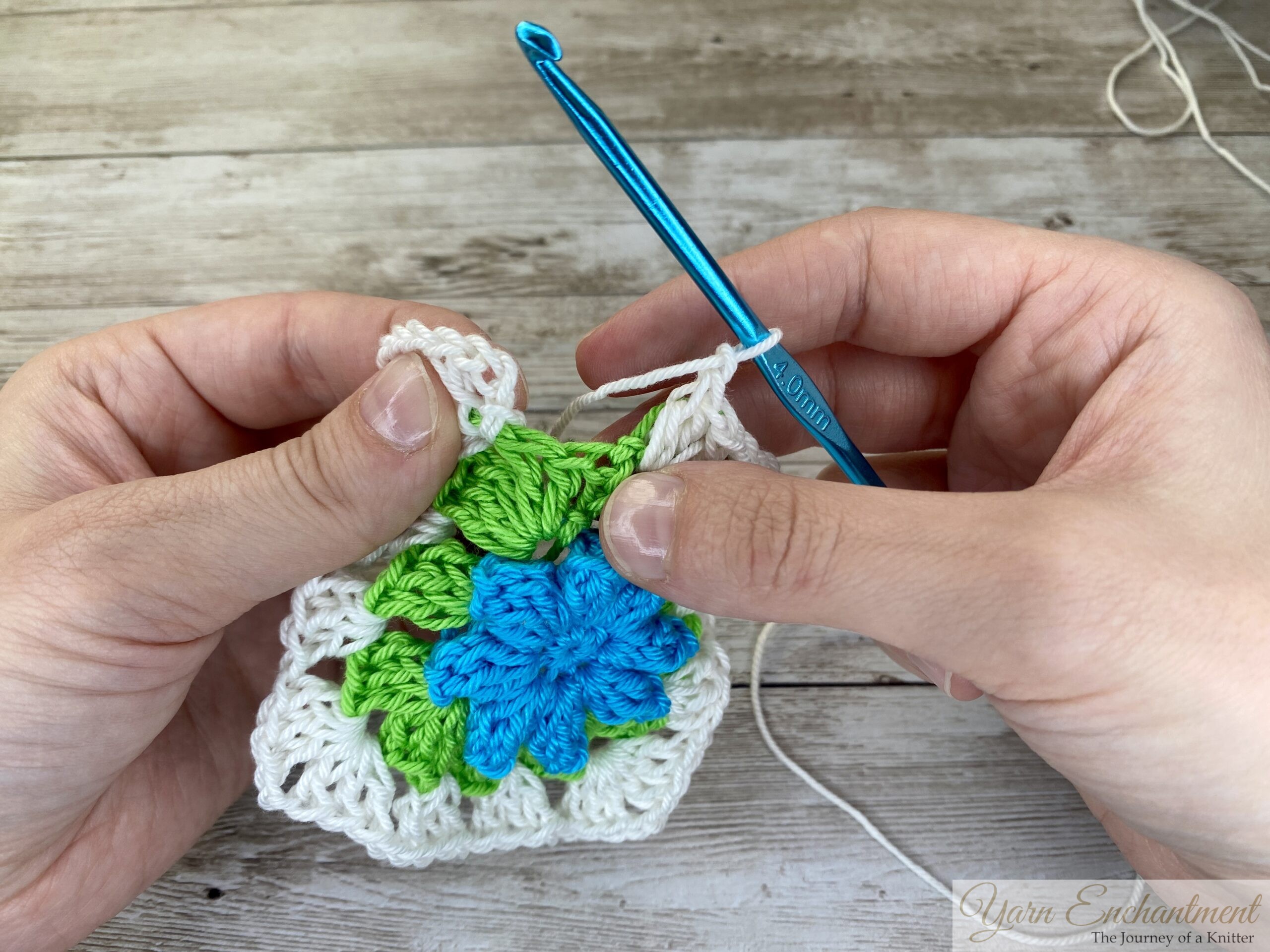The crochet project advances with white yarn forming a detailed border around the green and blue granny square. A blue crochet hook is held in position as the piece takes shape