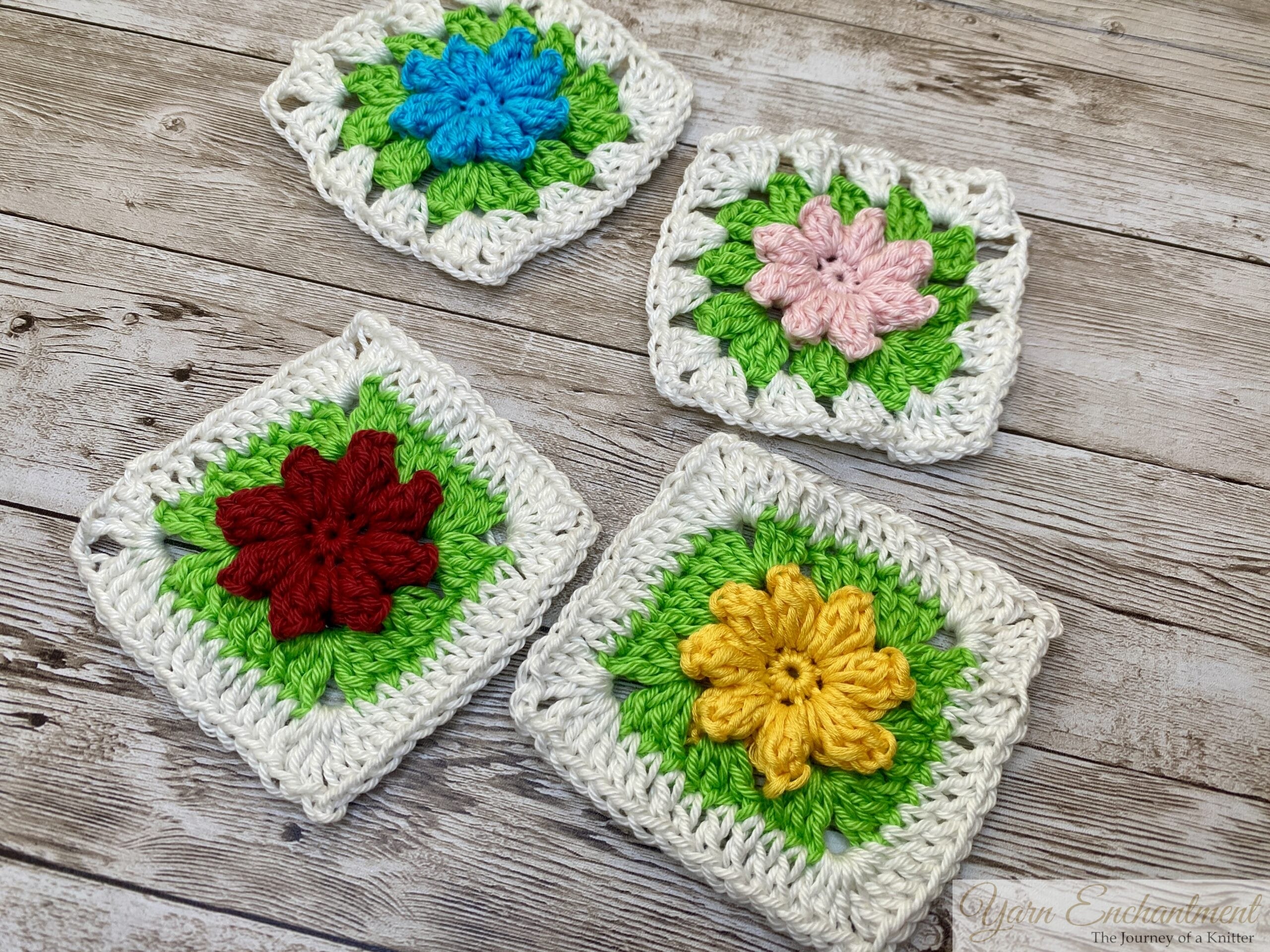 Angled view of four 3D flower granny squares on a wooden surface, showcasing vibrant flowers in blue, red, pink, and yellow, framed by green petals and white borders