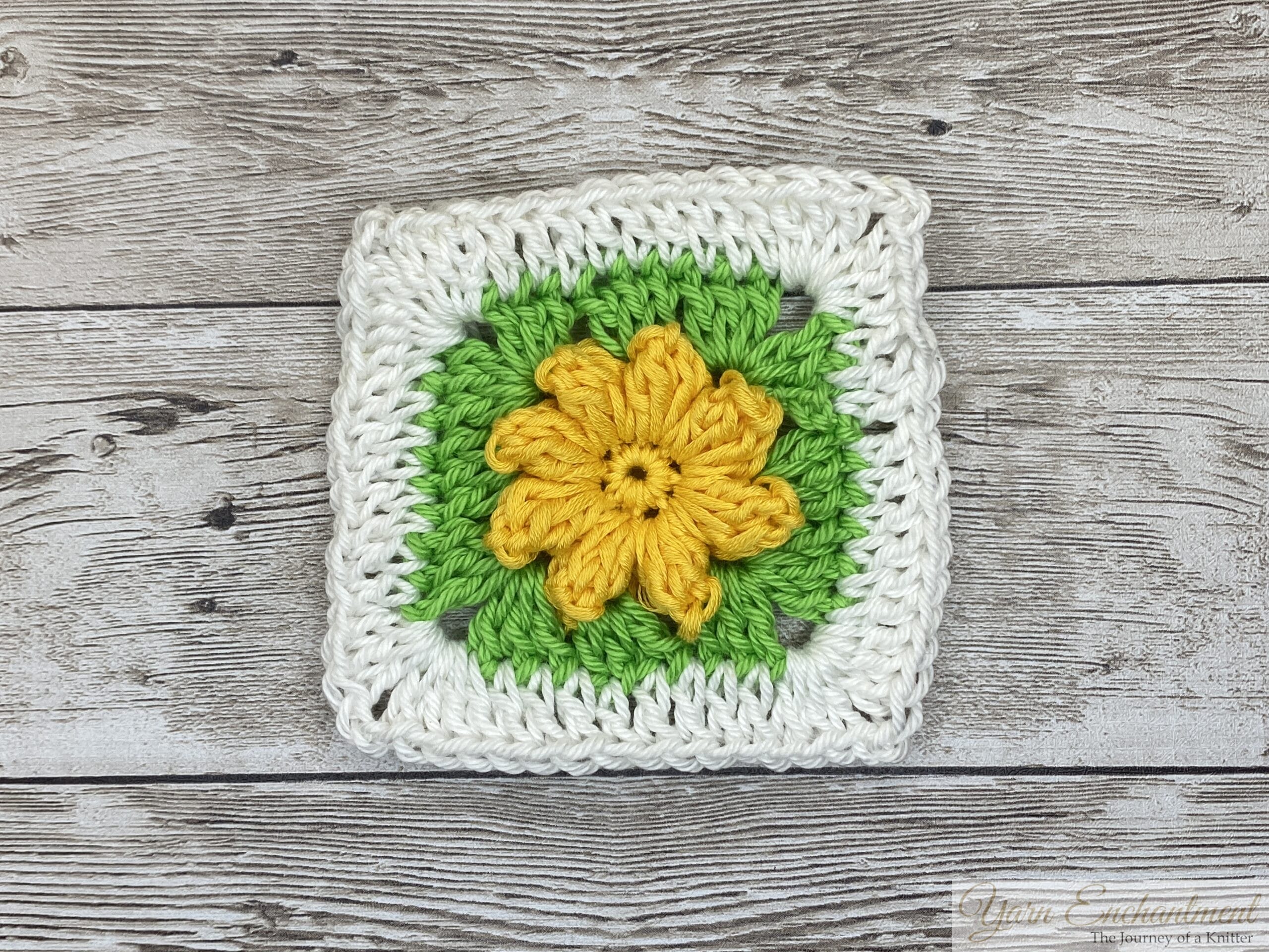 A single 3D flower granny square with a bright yellow flower centerpiece, framed by green petals and a white crocheted border, set on a wooden background.