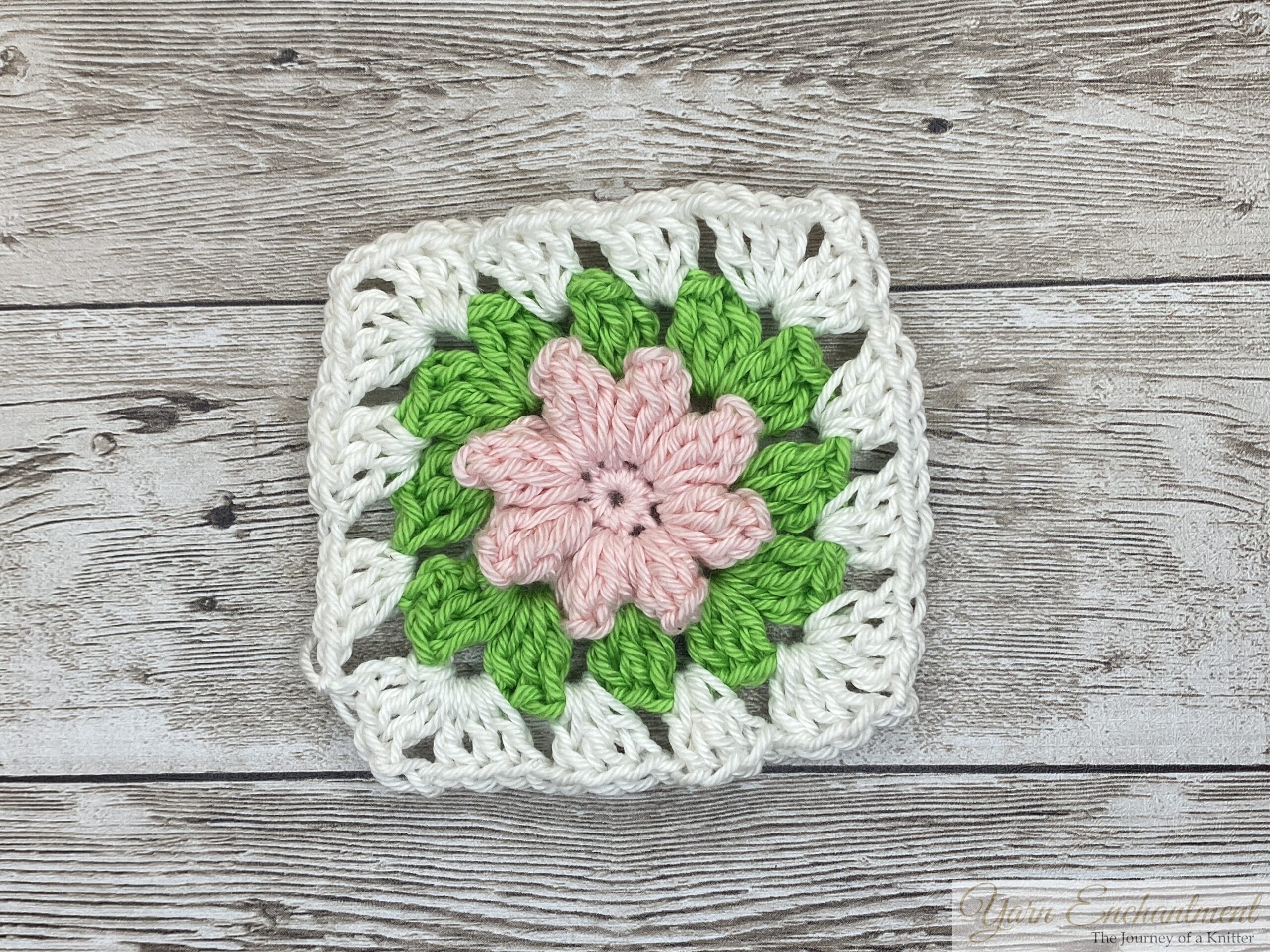 A single 3D flower granny square featuring a soft pink flower at the center, surrounded by vibrant green petals and a white border, displayed on a rustic wooden surface.