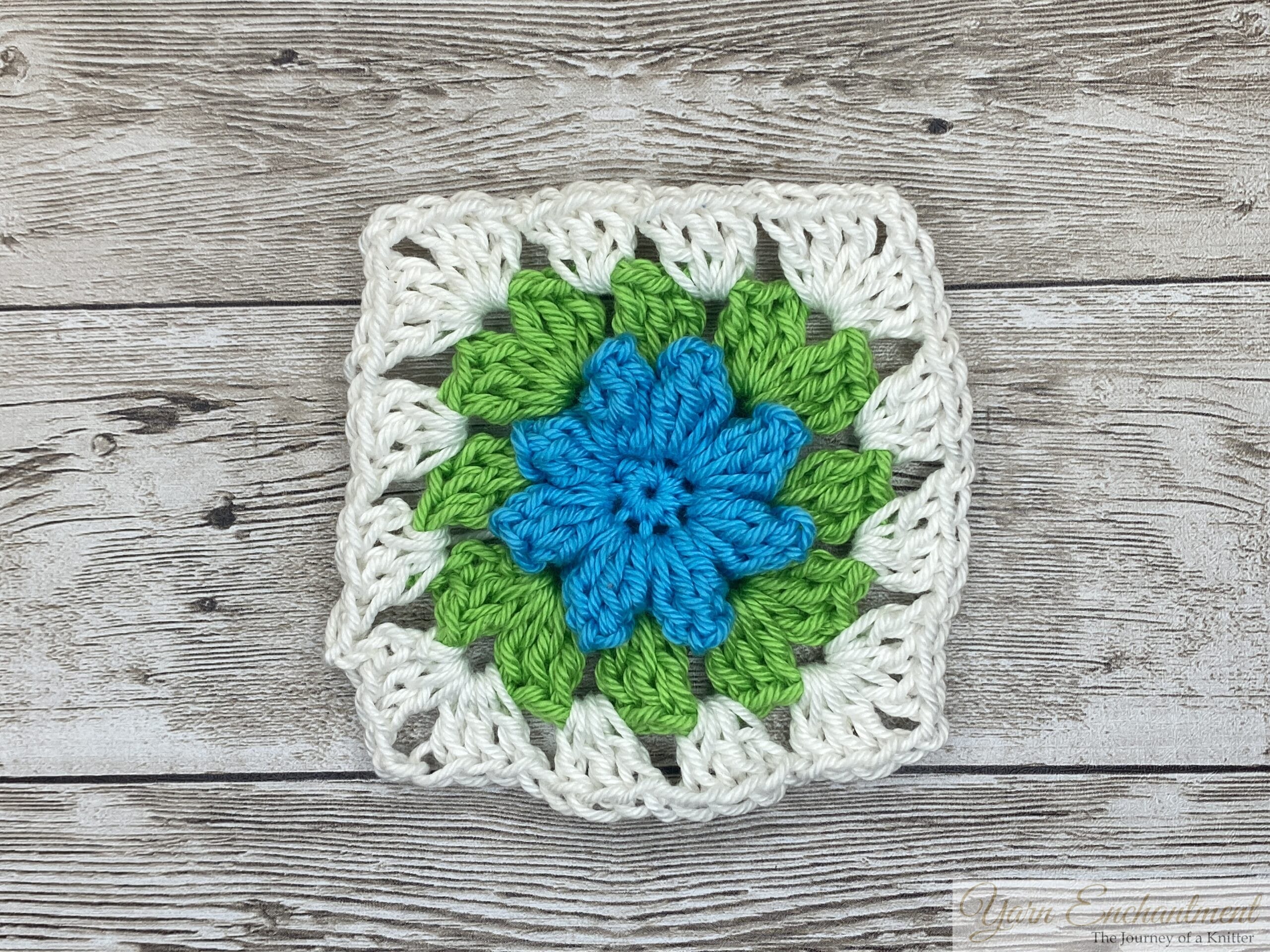 A single 3D flower granny square featuring a blue flower at the center, surrounded by vibrant green petals and a white border, displayed on a rustic wooden surface.