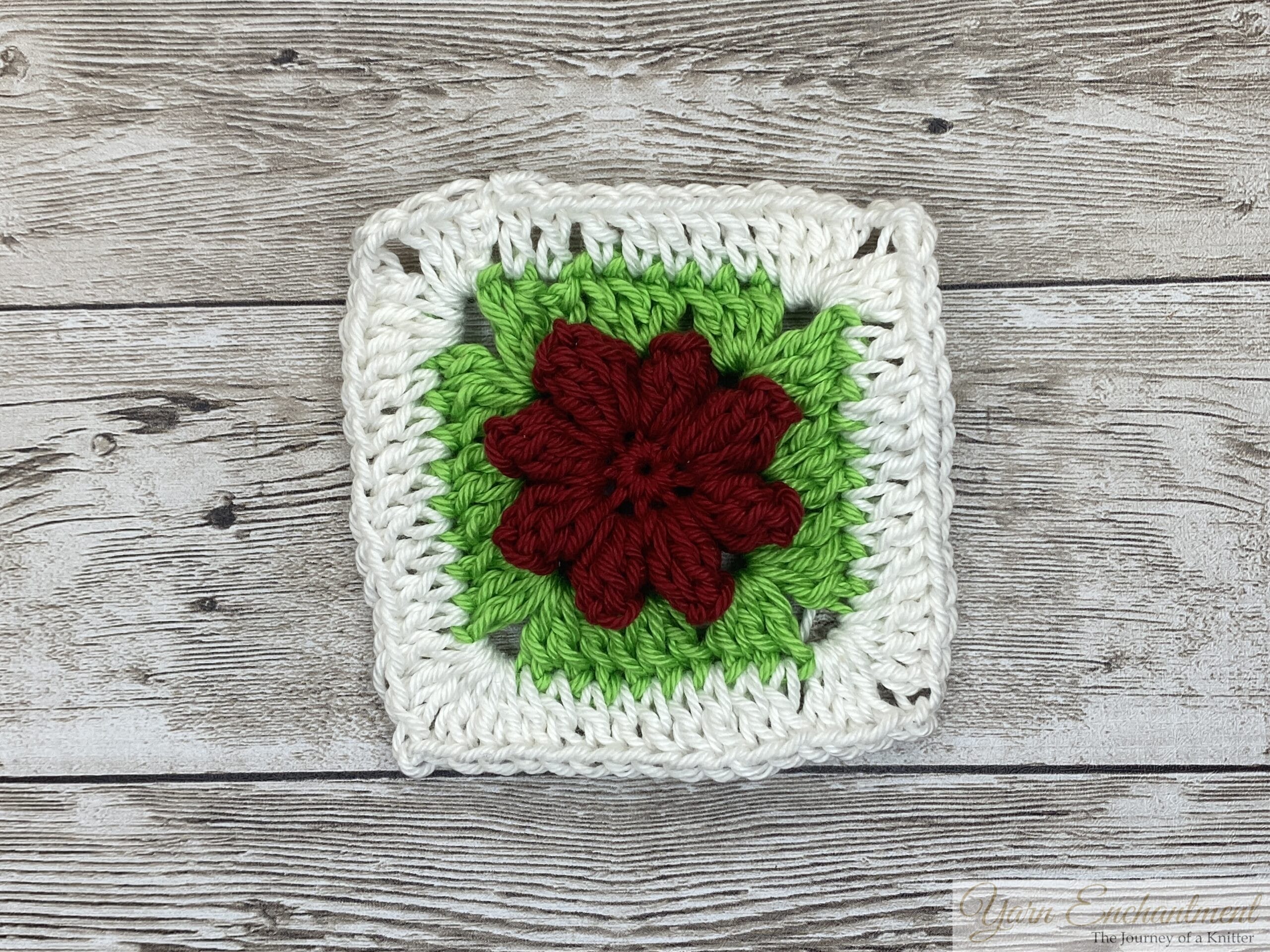 A single 3D flower granny square featuring a red flower at the center, surrounded by vibrant green petals and a white border, displayed on a rustic wooden surface.
