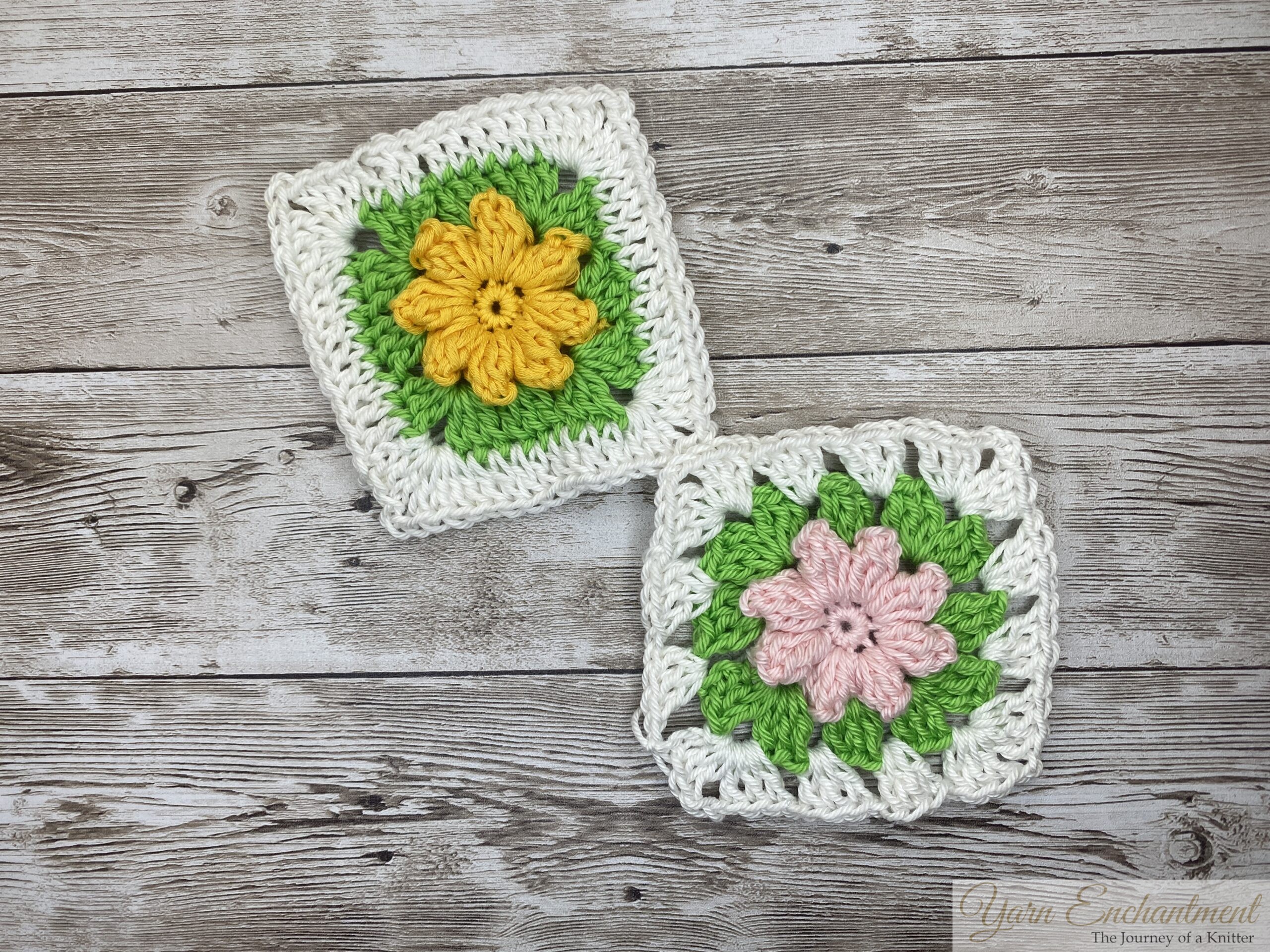 Two 3D flower granny squares, one with a bright yellow flower and the other with a soft pink flower, each surrounded by green petals and a white crocheted border, arranged on a wooden surface.