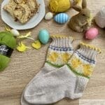 A pair of hand-knitted socks made with Drops Fabel and Drops Flora yarn, featuring a spring-themed colorwork design with yellow chicks, green grass, and blue accents. The socks use the Latvian Twist cast-on technique and have a decorative yellow scalloped trim. They are displayed on a wooden surface surrounded by Easter decorations, including painted eggs, yellow wooden chicks, balls of yarn, a ceramic bunny, and a plate of sliced bread. The arrangement creates a festive and cozy atmosphere.