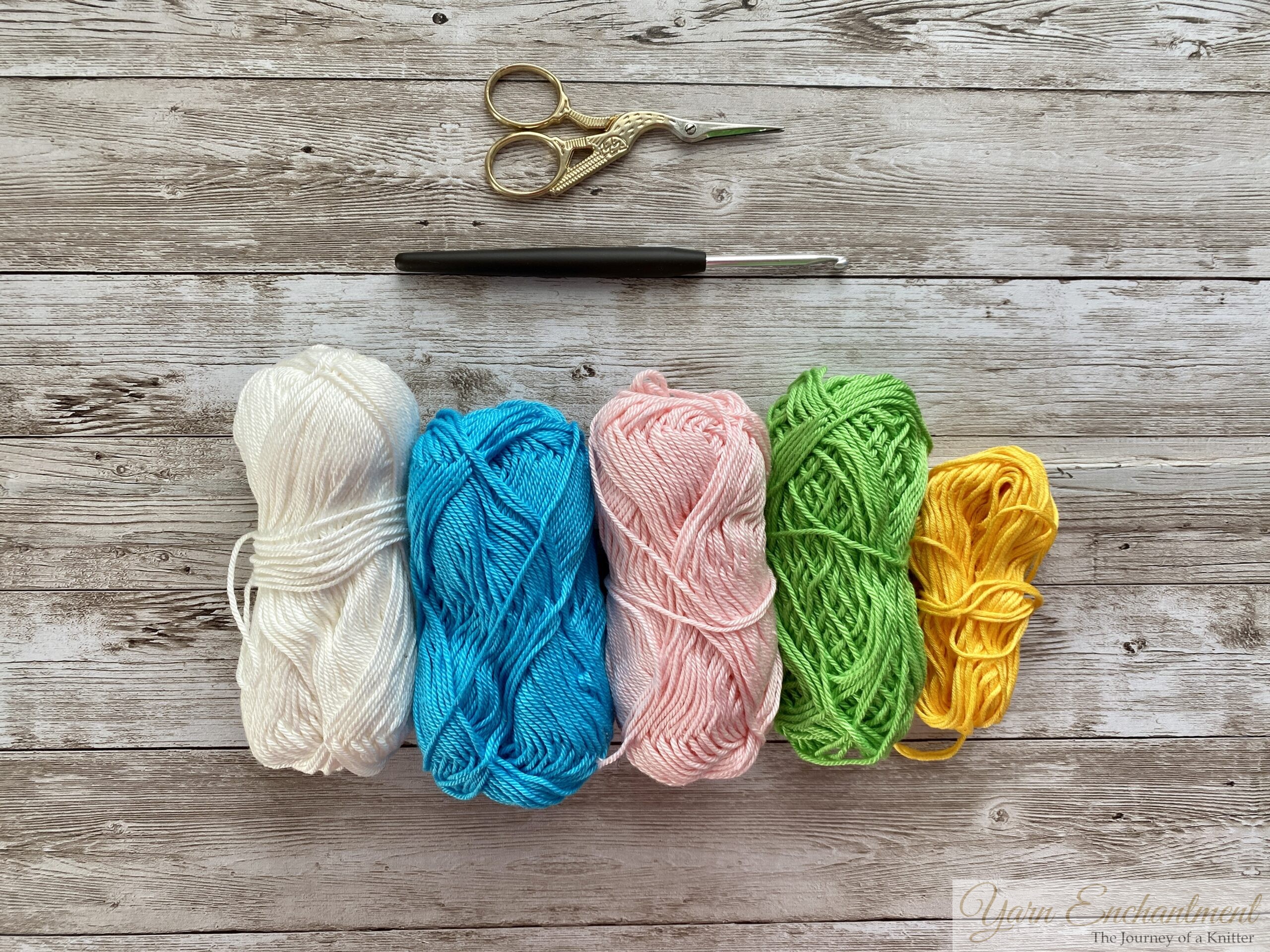 Assorted crochet materials laid out: a pair of scissors, a crochet hook, and skeins of yarn in white, blue, pink, green, and yellow for making 3D granny squares.